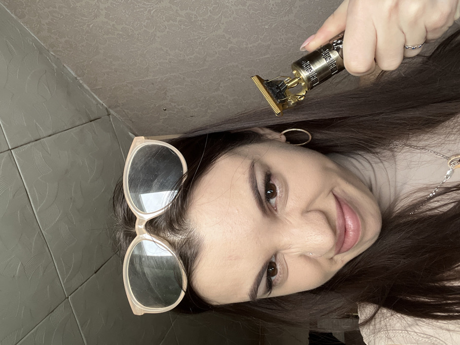 The image depicts a young adult female in an interior room within the city of Chaikovsky, Russia. The subject has long, dark brown hair, with light-colored, oval-framed eyeglasses resting on top of her head. She wears a light-colored, possibly beige, turtleneck garment and a silver-toned necklace. Her right ear features a gold-colored hoop earring, and a small stud is visible on her left nostril. Her facial expression is a playful grimace, with her nose slightly scrunched and her mouth downturned, as she looks directly towards the camera. She holds a metallic gold-colored electric hair trimmer in her right hand, which displays intricate etched patterns on its body. The background consists of a dark-toned wall with a subtle, repeating floral or damask pattern, meeting a ceiling composed of dark grey or black textured square panels.