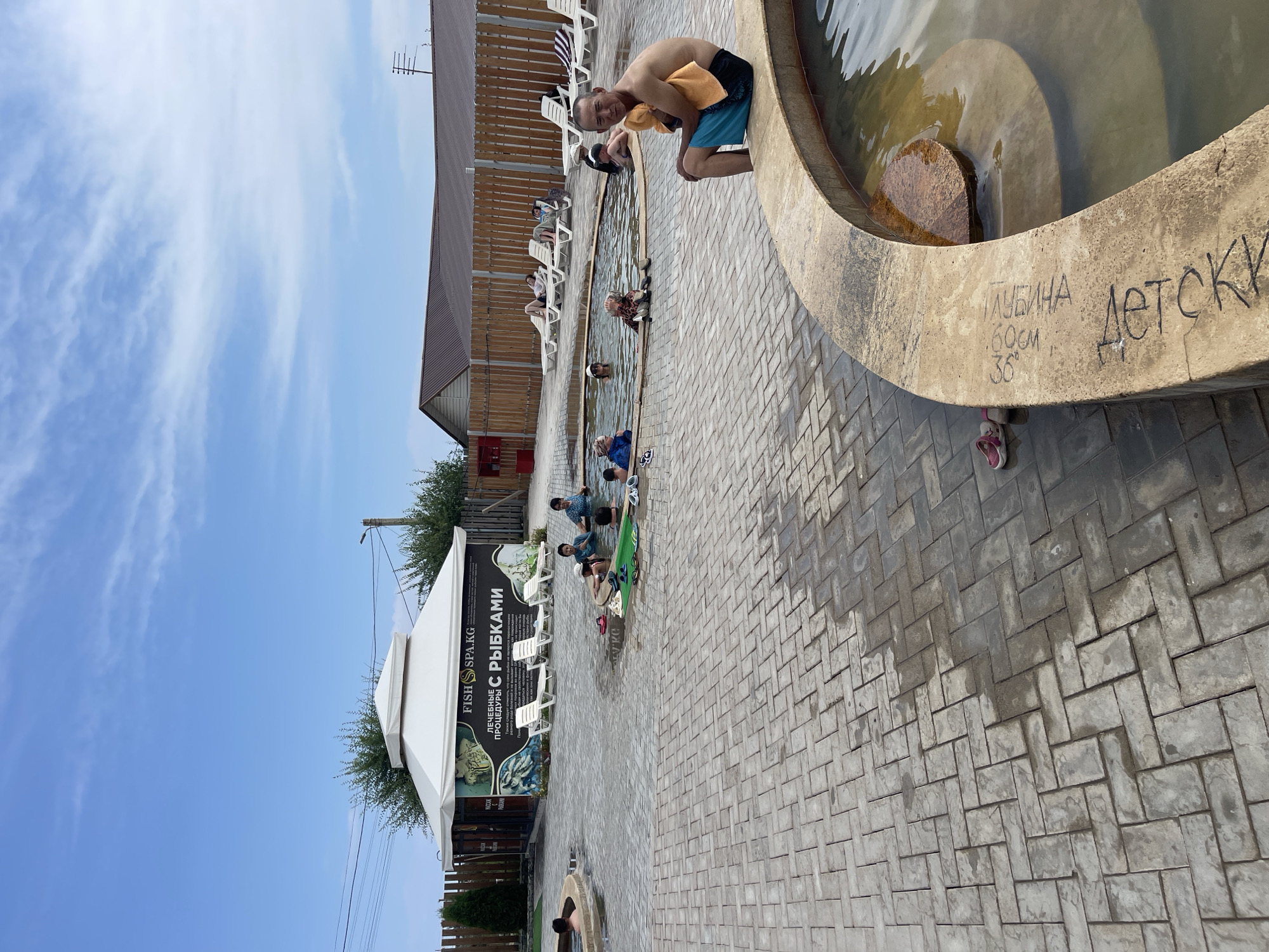 The image depicts an outdoor fish spa in Cholpon-Ata, Kyrgyzstan. Several people are visible submerged in warm water pools, some engaging in fish pedicures, while others relax on lounge chairs. A man in the foreground is seated on the edge of a pool, holding an orange towel. The area is paved with interlocking gray bricks, and a white canopy structure with signage for "FISH SPA.KG" is present. The environment appears to be a recreational facility with multiple pools of varying sizes and depths. The sky is partly cloudy.