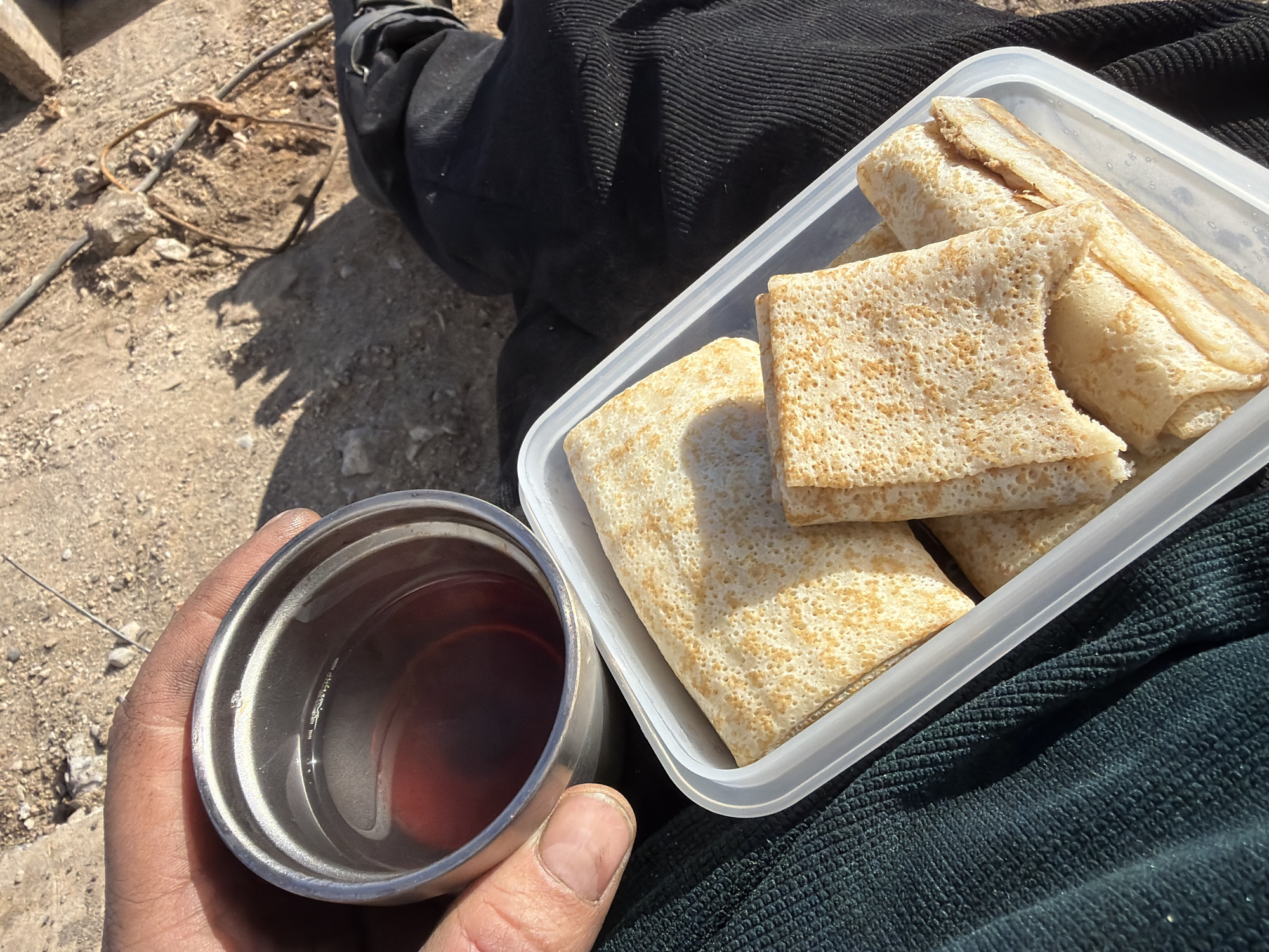 A person is having a meal outdoors. They are holding a metal cup filled with a reddish liquid. Next to the cup is a clear plastic container holding several rolled pancakes. The person is wearing dark clothing. The ground is visible in the background and appears to be dirt with some gravel and debris. Based on the location information provided, this scene is taking place in Chaikovsky, Russia.