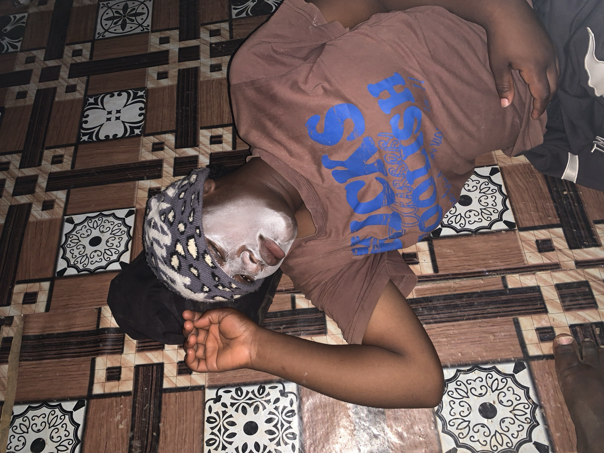 A person lies on a tiled floor. The person's face is covered in a white substance. The person wears a brown t-shirt with blue lettering, and a patterned head covering. The setting appears to be indoors, based on the flooring. Part of one foot is visible in the lower right corner. No information on location in Agban, Nigeria, is available.