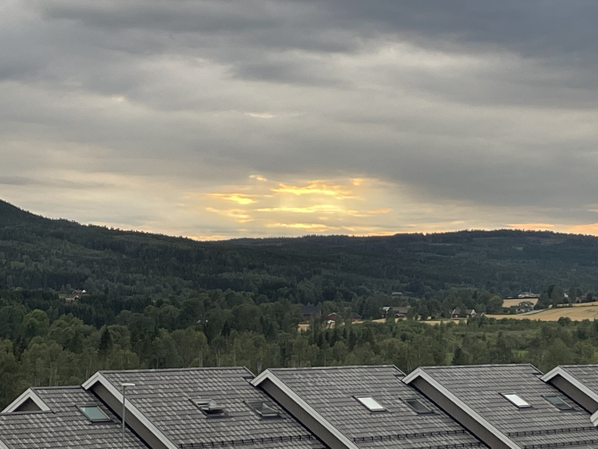 This is a wide shot of a Norwegian landscape in Løvstad, Norway. The foreground is dominated by the dark grey tiled roofs of several modern houses, each with visible skylights. Beyond the houses, a vast, dark green forest covers rolling hills, with scattered red-roofed buildings visible within the tree line. In the distance, a lighter green field contrasts with the forest. The sky is filled with dramatic, grey clouds, but a break in the clouds reveals a bright, sunlit area where the sun's rays are visible. The overall impression is of a tranquil, slightly overcast, rural setting.