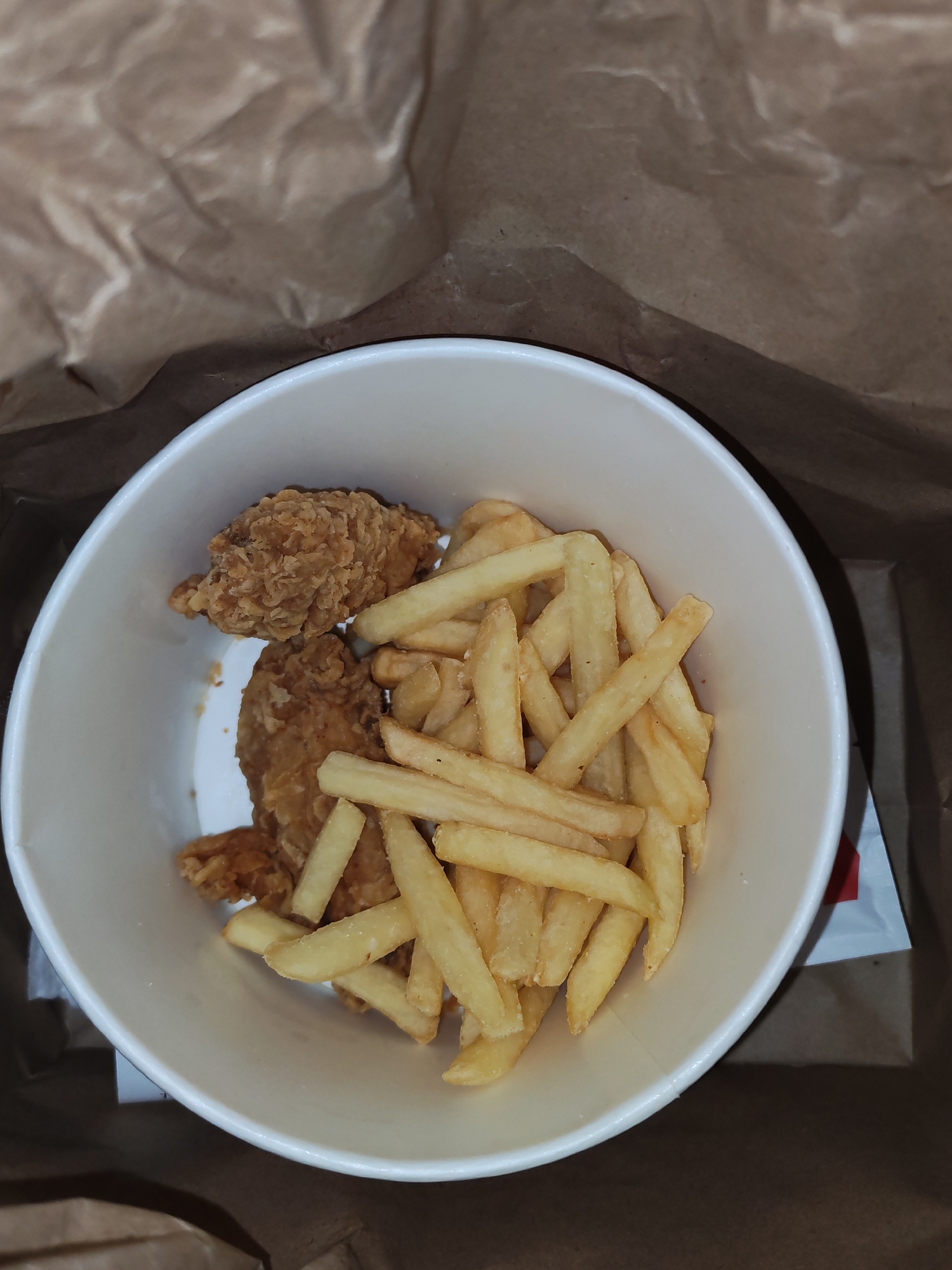 A top-down, close-up shot shows a white paper bucket filled with fried chicken pieces and french fries. The bucket is partially submerged in a brown paper bag, which is crumpled and textured. The fried chicken pieces are a golden brown color and appear to have a crispy coating. The french fries are cut into thin strips and are also golden brown, some showing slightly darker, crisped edges. The scene is likely an indoor setting, possibly a restaurant or home, and the image is taken at an angle that emphasizes the food within the bucket. There is no indication of the specific location in Izhevsk, Russia, other than what is described.