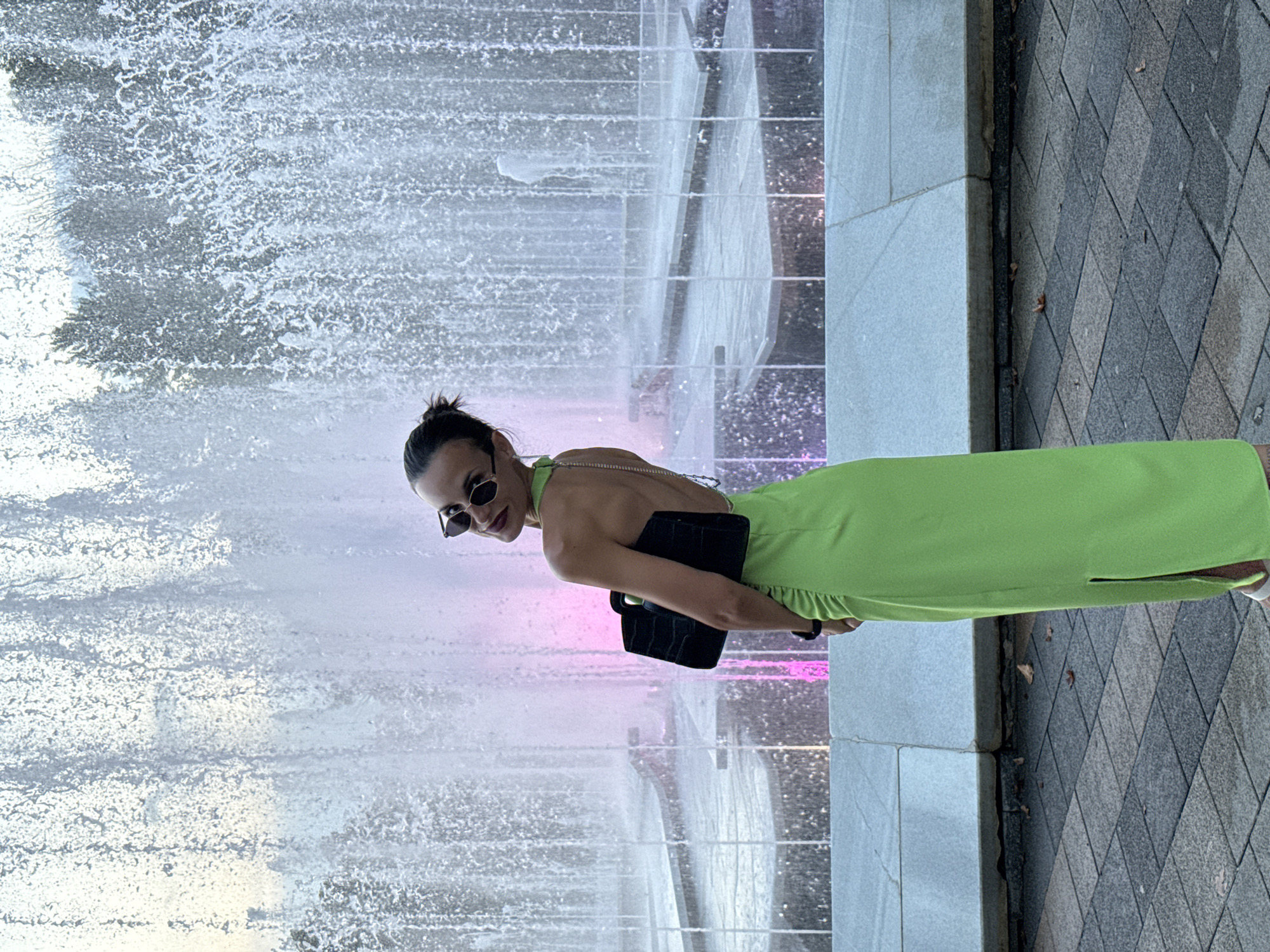 A woman is posing by a fountain in Pleven, Bulgaria. She is wearing a lime green, backless dress with a side slit, sunglasses, and carrying a black handbag. The fountain's water is illuminated with pink and white lights, creating a vibrant backdrop. The ground is paved with gray and black square tiles.