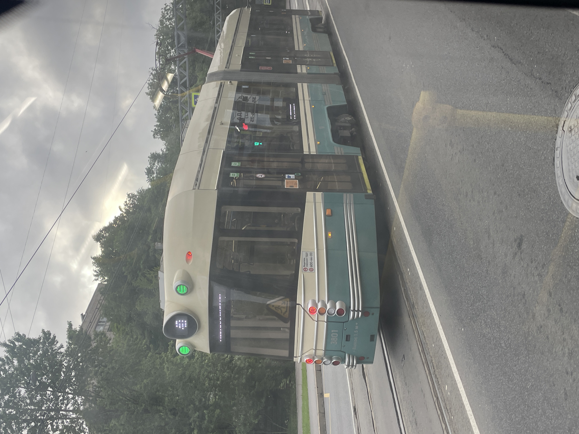 The image shows a modern tram in Saint Petersburg, Russia, traveling on a street with tram tracks. The tram is cream and teal, with multiple windows and doors. It has illuminated headlights and taillights. The tram has the number "3801" visible on its rear. The background consists of trees and a cloudy sky. A yellow traffic bollard is visible in the foreground on the right side of the image. The scene appears to be captured from another vehicle, likely a car or another tram, given the reflection on the glass in the foreground.
