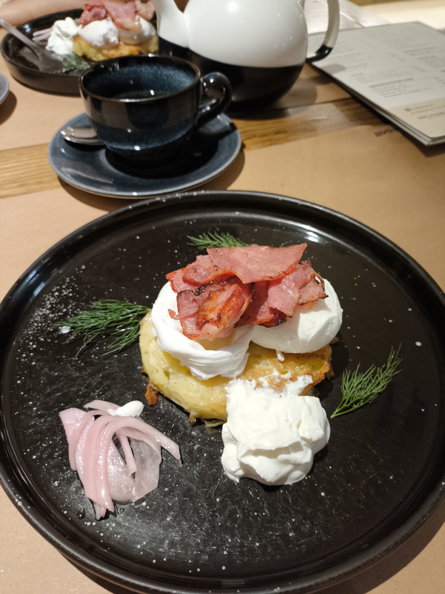 The image depicts a meal served on a wooden table within an indoor setting. In the foreground, a round, black ceramic plate with white speckles holds a savory dish. The central item is a stack consisting of a golden-brown base (likely a potato pancake or fritter), topped with two mounds of white cream or poached egg, and crowned with several slices of reddish-pink, cooked bacon or cured meat showing some crisp edges. To the lower left of the main dish, there is a small pile of thin, pale pink, pickled onion slices, partially covered by a small dollop of white cream. To the lower right, a larger dollop of white, thick cream or sour cream is present. Two small sprigs of fresh green dill serve as garnish, positioned on the left and right sides of the main food stack. White granular specks, possibly salt, are scattered on the left side of the black plate.
In the mid-ground, to the left, another partially visible dark plate holds a similar food item, including a golden base, white topping, and meat, with a silver utensil resting on it. Directly behind the foreground plate, a dark blue or teal ceramic cup with a textured finish, containing a dark liquid, sits on a matching saucer with a silver spoon. To the right, a white ceramic teapot or pitcher with a black handle and base is partially visible. Further right, a stack of paper, appearing to be a menu or leaflet with text in Latin script, rests on the table. The table surface consists of light brown wooden planks, partially covered by a lighter, possibly paper or fabric, surface under the plates. No individuals are visible, and no specific actions are being performed. The lighting is warm and indirect. The location in Izhevsk, Russia, cannot be determined from the visual content of the image.