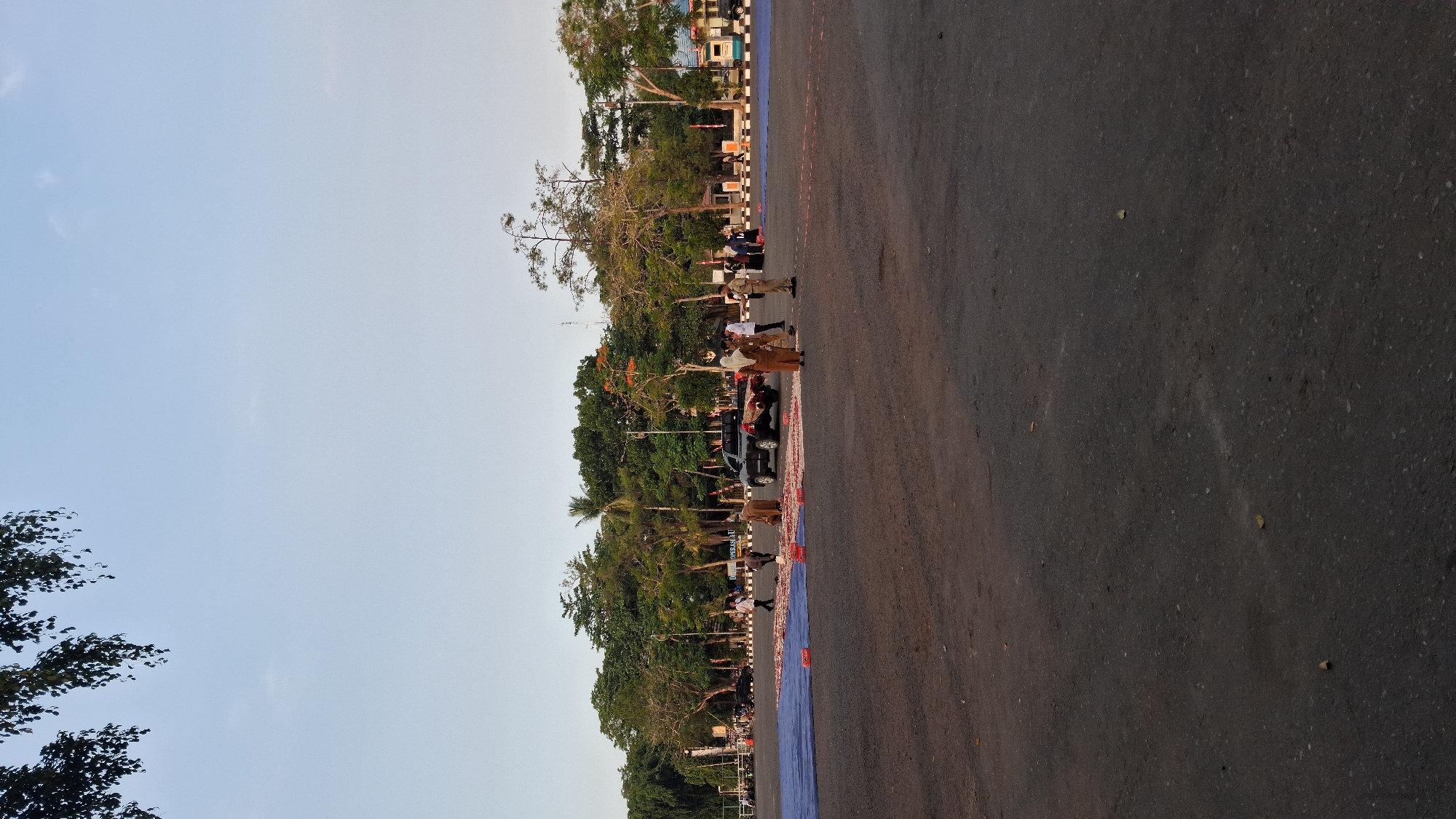 The image depicts an outdoor scene in Banjarbaru, Indonesia, on an asphalt-paved area, likely a parking lot or an open field, with a backdrop of lush green trees and a clear sky. In the foreground, several individuals are gathered around a dark-colored pickup truck and a large blue tarp spread on the ground. Some people are wearing traditional Indonesian attire, while others are in uniforms. The blue tarp is adorned with a scattering of red and white objects, possibly flower petals or confetti, arranged in a pattern. A black SUV is parked near the truck, and more red and white objects are visible on the ground in front of it and extending towards the right side of the frame. The area appears to be prepared for an event or ceremony, with the arrangement of the tarp and the scattered items suggesting a celebratory or commemorative purpose. The presence of trees suggests the location is in a relatively urban or semi-urban area within Banjarbaru.