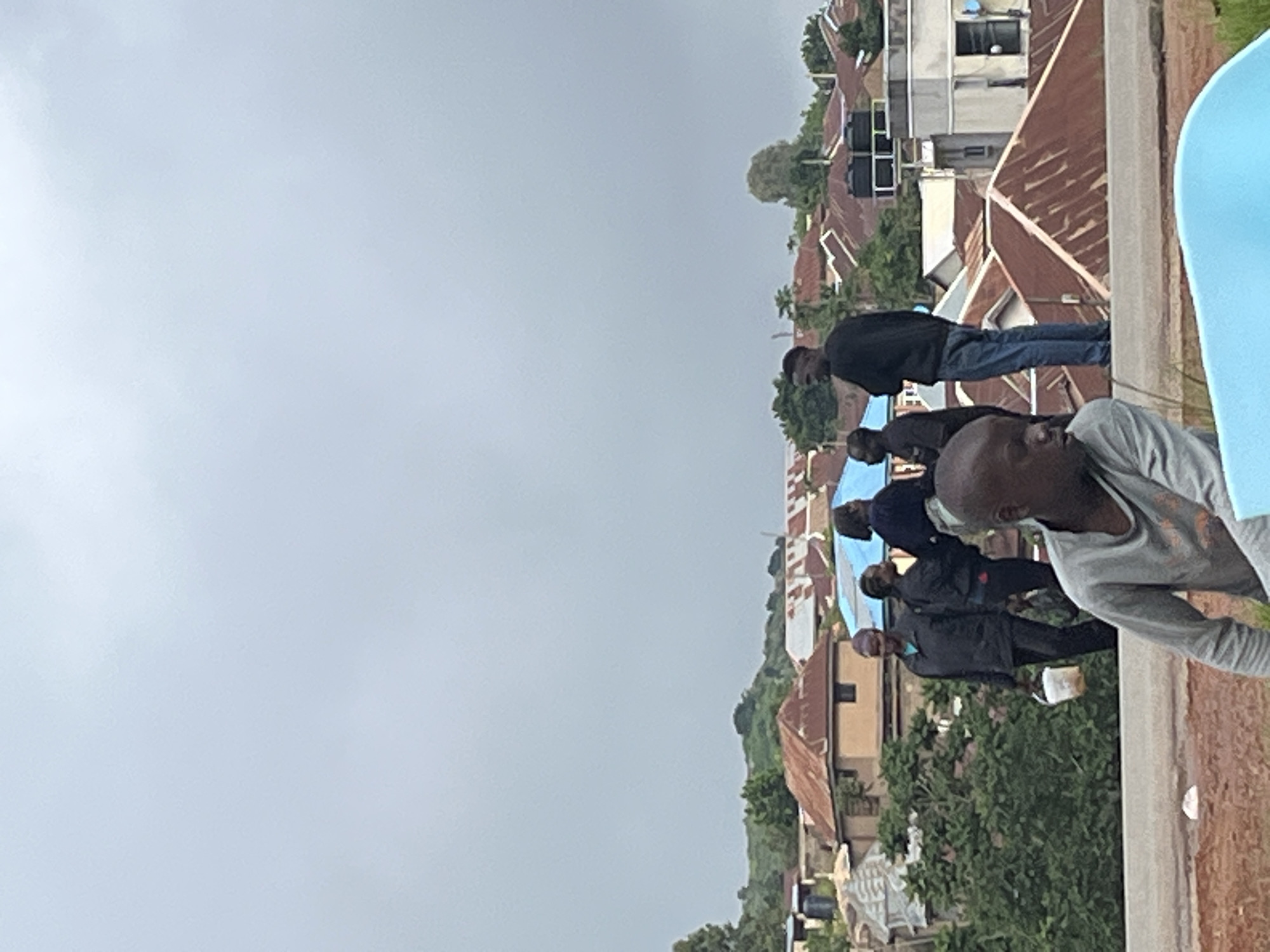 In Jos, Nigeria, on a cloudy day, several people are gathered near a cluster of residential buildings with corrugated metal roofs. In the foreground, a young man wearing a gray shirt looks down. To his right, a portion of a light blue object, possibly a tub, is visible. Behind him, a group of individuals stand on a rooftop. Among them, a woman in a black jacket carries a white bucket. Another man in a black shirt and blue jeans stands with his arms crossed. The background reveals houses and lush green trees, indicating a residential area.