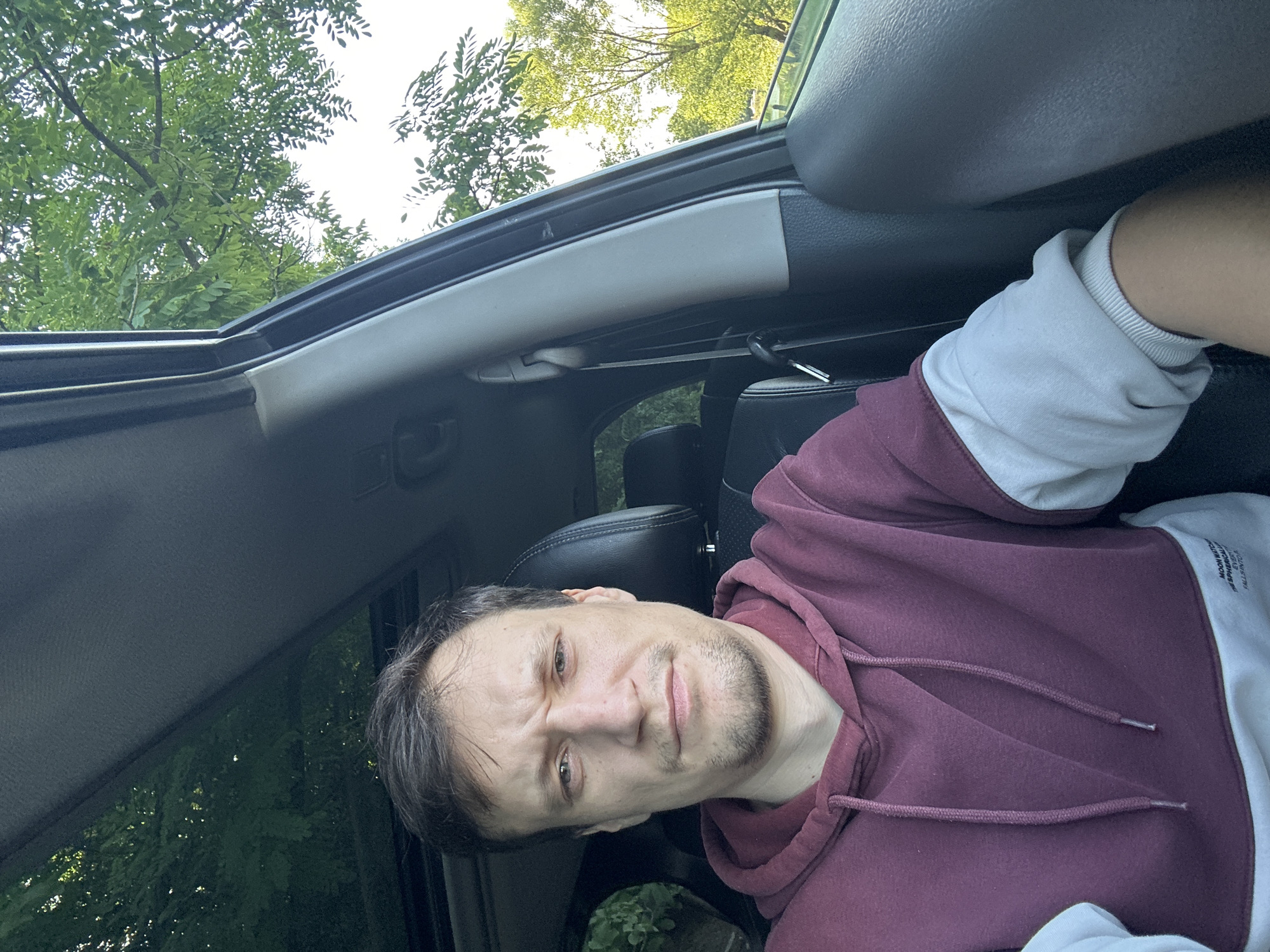 A man is taking a selfie from the driver's seat of a car. He is wearing a maroon and white hoodie. He has short, dark hair and is looking at the camera with a neutral expression. The car's sunroof is open, and green trees are visible through it, indicating an outdoor setting. The man's left arm is resting on the car's armrest, and the interior of the car, including the dashboard and headrest, is visible. The exterior environment suggests a rural or suburban area with lush greenery. The specific location in the city of Makiv, Ukraine cannot be determined from the image alone.