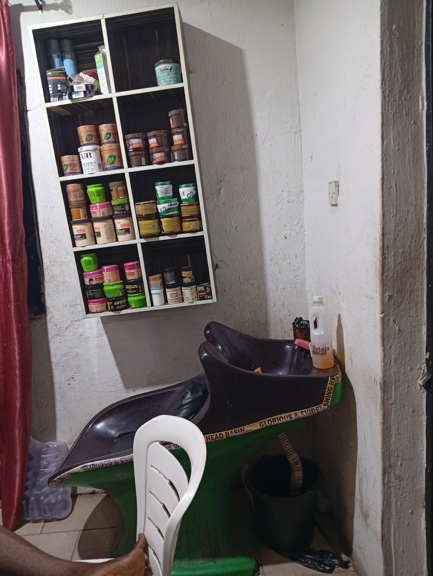 The image depicts the interior of what appears to be a small hair salon or beauty shop. A hair-washing station is visible, consisting of a dark-colored basin with a green stand that has writing on it, a white plastic chair next to it, and a black bucket placed below the basin to catch water. A bottle labeled "Petals Conditioner" sits on the edge of the basin.
A white shelf unit mounted on the wall contains various hair products, including jars and bottles. The products display brand names. A red curtain is visible on the left side of the image. The walls are painted white. The floor is tiled. A partially visible person is in the lower left corner of the photo. The setting suggests a beauty establishment likely located in Lokoja, Nigeria.