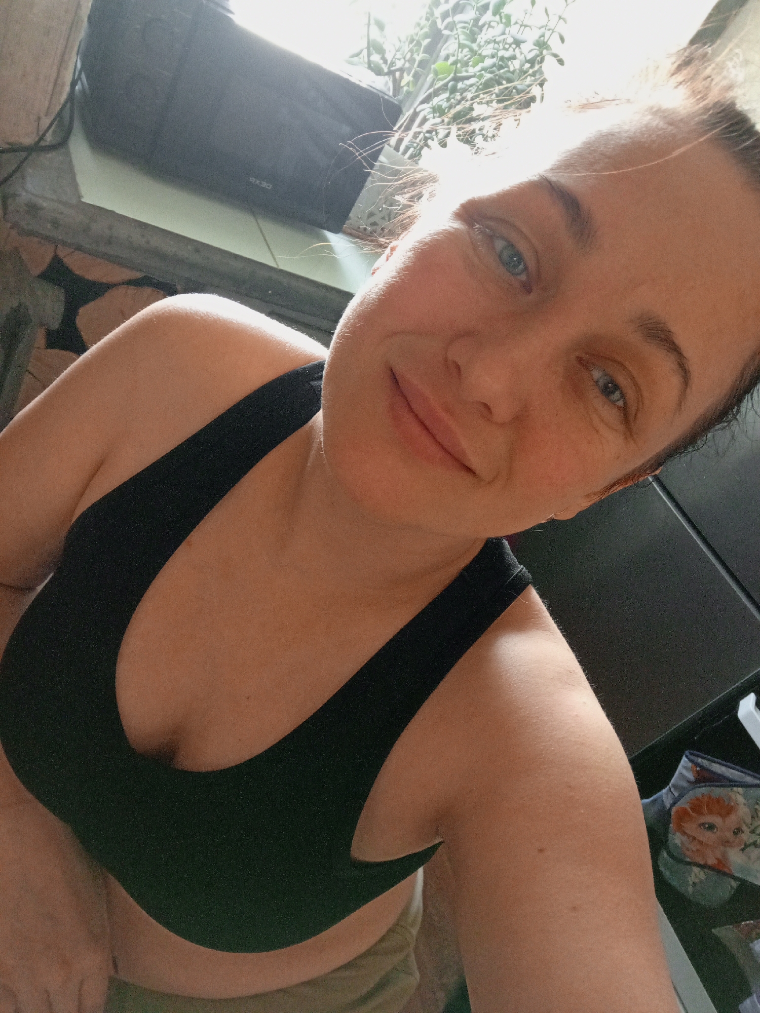 A fair-skinned woman with blue eyes and her hair pulled back is posing for a selfie indoors. She is wearing a black sports bra. Behind her is a wall with decorative wood and a black electronic device on a surface. In the upper right corner, there are green leafy plants visible. To the right, a portion of a black refrigerator can be seen, and there is a bag with a cartoon character next to it. The location is likely indoors, but the specific location in Chaikovsky, Russia cannot be determined from the image.
