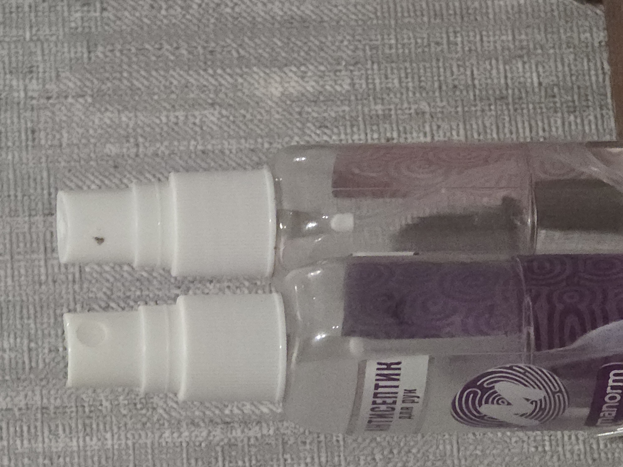 The image shows a close-up of two spray bottles against a textured, gray background. The bottle on the left is partially transparent and contains a purple liquid. A white label is attached to the bottle with Russian text that reads "АНТИСЕПТИК" and "ДЛЯ РУК" (Antiseptic for hands). Below the text is a circular logo with a stylized "M" inside concentric circles. The bottle on the right is also transparent and contains a light pink liquid with a pink patterned label. Both bottles have white spray nozzles. The scene is indoors and appears to be a still life composition. There are no people or animals visible in the image.