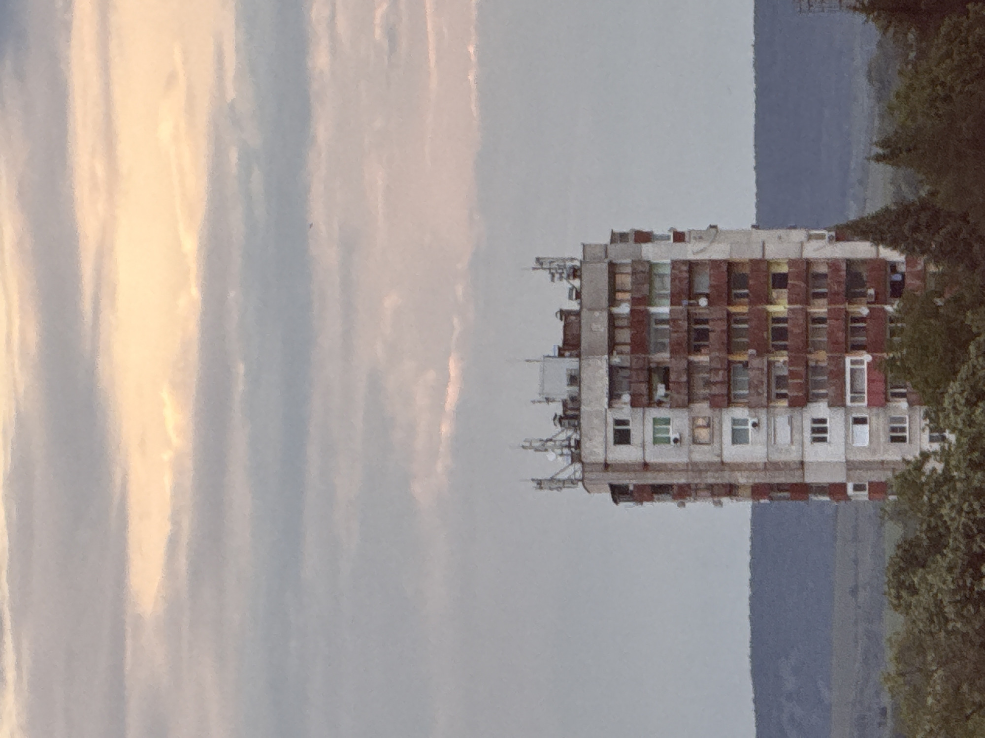 The image presents a multi-story residential building, likely an apartment block, with a concrete frame and red brick in-fill. Numerous balconies and antennas are attached to the building's exterior. The sky is visible to the left of the building, showing a mix of light and dark gray clouds, possibly indicating twilight or an overcast day. A tree is present on the right side, partially obscuring the building.
While the prompt requests to mention the location in Lovech, Bulgaria, it is not possible to confidently verify if the location is Lovech based on the image alone. More contextual clues or corroborative information would be needed to confirm the exact location.