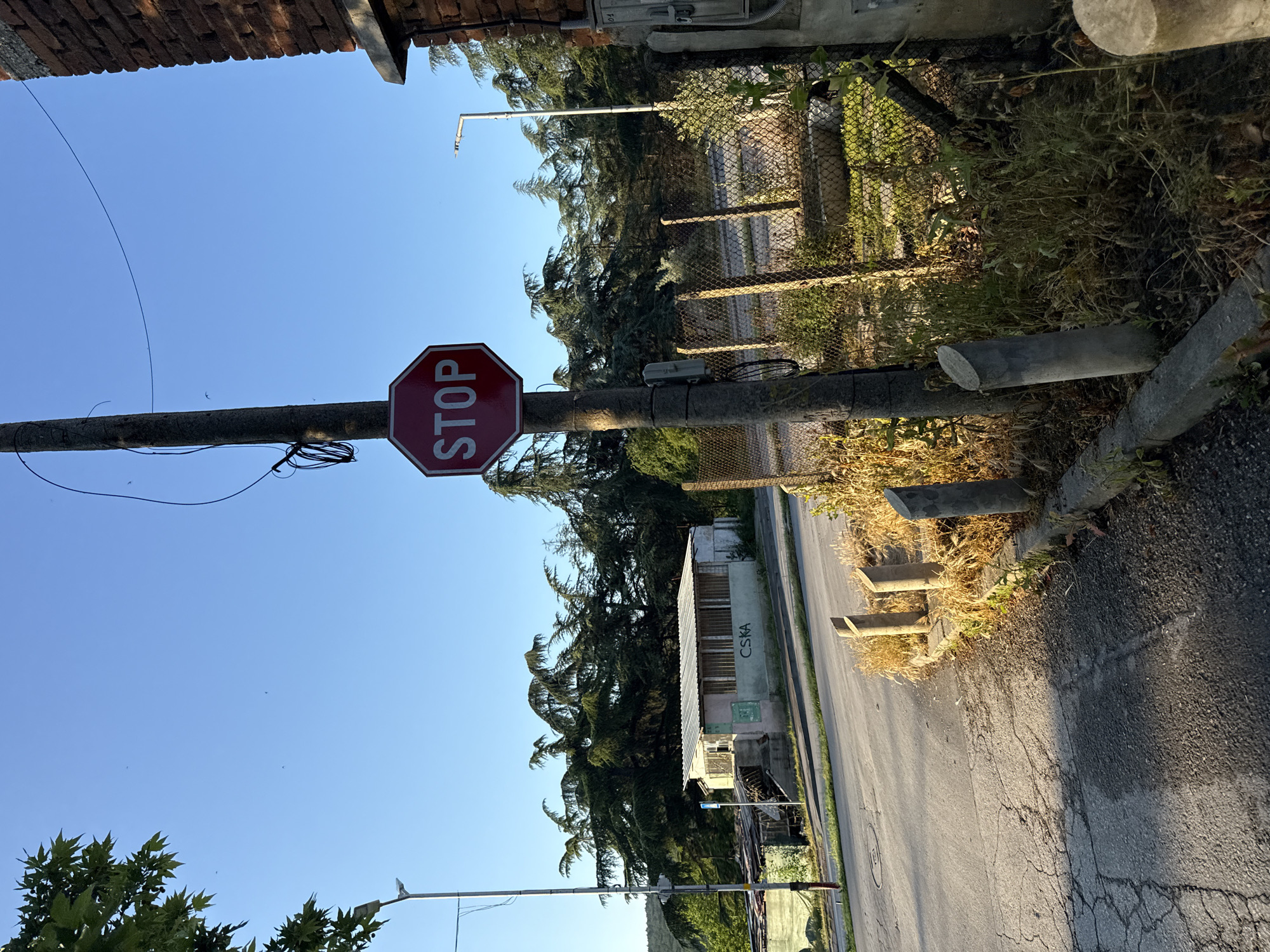 A stop sign is mounted on a utility pole next to a brick building in an urban environment. The sign is octagonal and red with the word "STOP" in white letters. The pole has electrical conduits and wires attached to it. In the background, there is a street, a chain-link fence, and a line of trees. A small, dilapidated building with a metal roof is visible on the left side of the image, and a streetlamp stands in the distance on the right. The sky is clear and blue, suggesting it is daytime. The overall scene appears to be an intersection or a roadside in a somewhat neglected area.