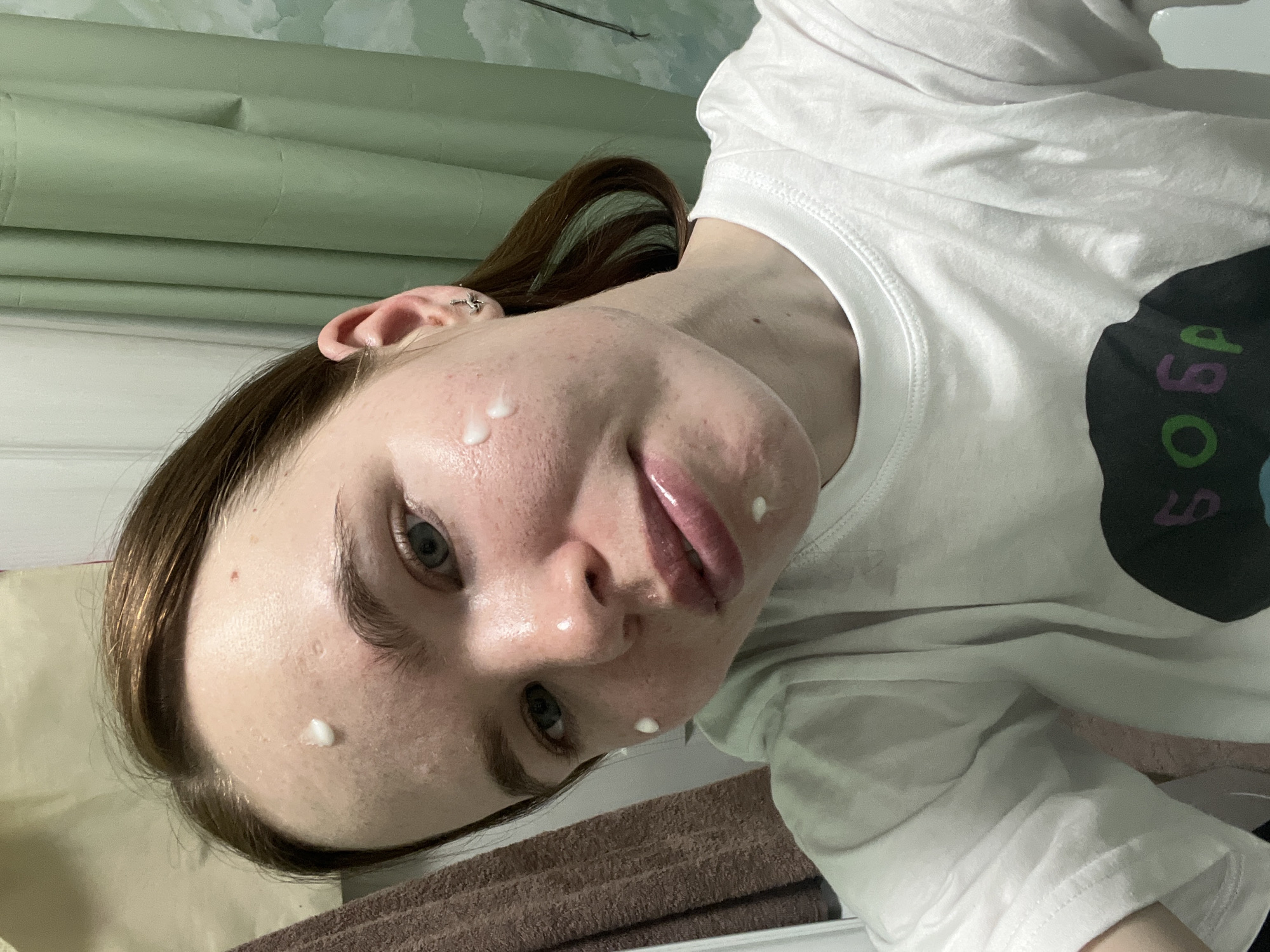 A young woman is pictured indoors, likely in a bathroom, as indicated by the pale green patterned wall and partial view of what appears to be a brown towel. She has fair skin, light blue eyes, and brown hair with teal streaks. Her face is tilted toward the camera, and several small dollops of a white substance, possibly cream or lotion, are visible on her forehead, cheek, and chin. She is wearing a white t-shirt with a circular black design on the right side, featuring illegible green and purple text. The location of this photo cannot be reliably determined as Chaikovsky, Russia.
