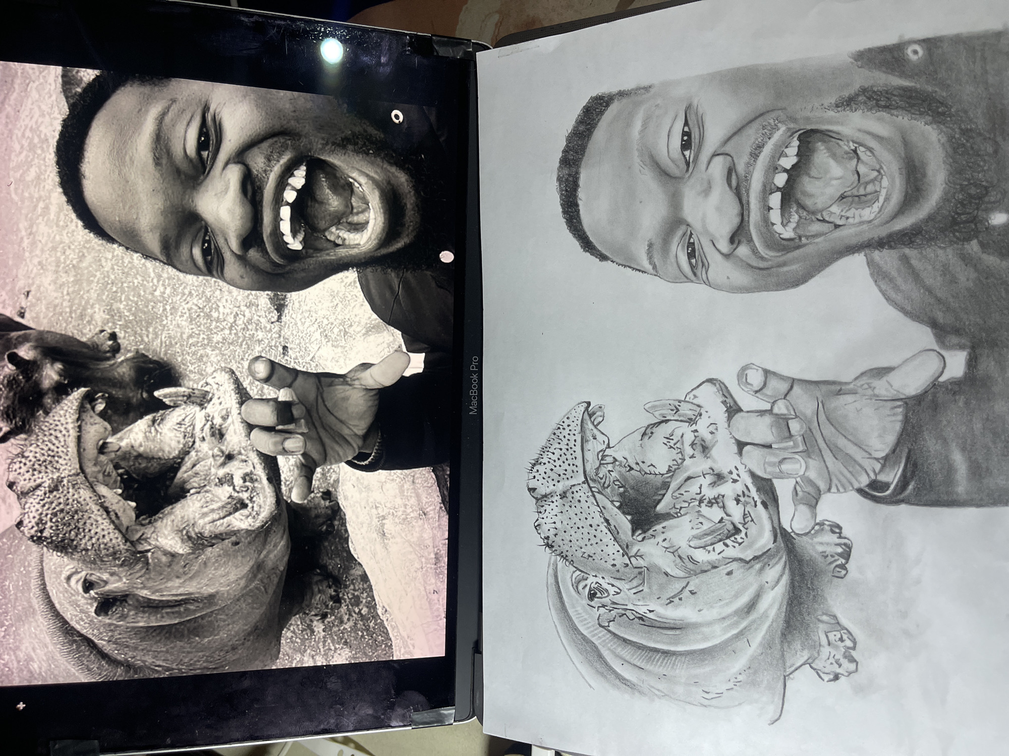 The scene displays an art workstation, centered on a silver MacBook Pro laptop open to reveal its screen, positioned above a sheet of white paper holding a graphite pencil drawing. This setting is located in the city of Jos, Nigeria.
The laptop screen presents a black and white photograph. On the left side of the photograph is an adult hippopotamus with its mouth wide open, exposing its large teeth, gums, and tongue. Its skin appears textured, and a portion of another animal is visible in the upper right background of the photograph. To the right, an adult male, likely of African descent, is depicted laughing exuberantly, with his mouth wide open, revealing visible teeth, some of which are gapped or missing. He has short, dark hair and a beard. His right hand is raised, partially obscuring the hippo's image, with fingers splayed. Both subjects in the photograph appear to be in an outdoor environment, possibly a wildlife enclosure or zoo setting.
Directly below the laptop, the white paper displays a detailed graphite pencil drawing that meticulously replicates the composition of the photograph on the screen. The drawing features the same wide-mouthed hippopotamus on the left and the laughing man on the right, rendered with accurate shading and texture to convey realism. The man's facial expression, hair, beard, and raised hand, along with the hippo's features, are accurately reproduced in the drawing. The laptop's base is dark, with "MacBook Pro" branding visible. The overall arrangement illustrates the artistic process of using a digital reference image for a traditional drawing.