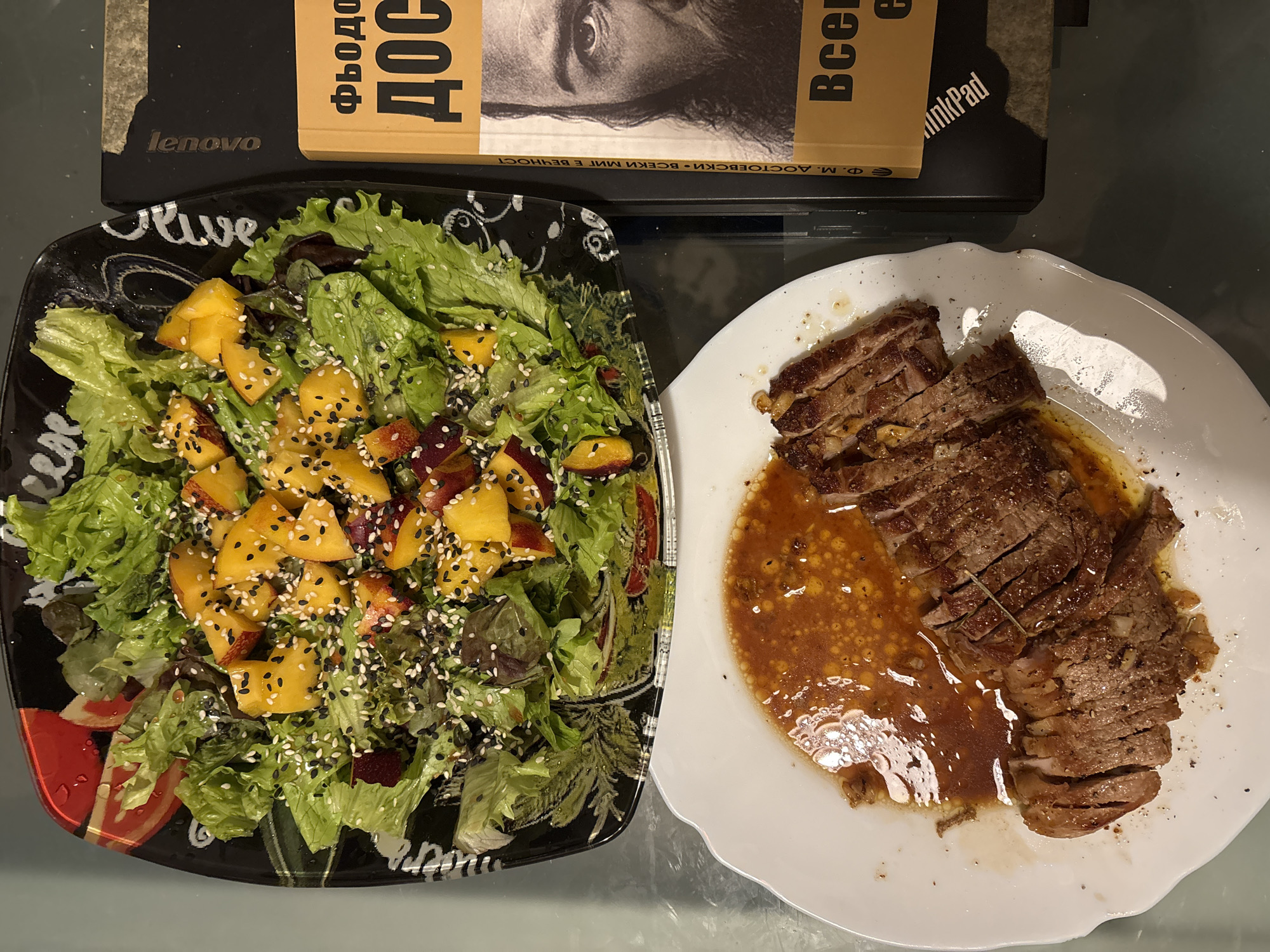 A close-up, top-down shot captures a meal laid out on a reflective, dark grey surface, likely a table. To the left, a black, ornately patterned salad bowl holds a vibrant green salad adorned with yellow peach slices, dark red beet pieces, and a scattering of white and black sesame seeds. The salad appears to be dressed with a light vinaigrette.
Below the salad bowl, a white, scalloped-edge plate displays sliced, medium-rare steak. The steak is fanned out and coated in a glistening, reddish-orange sauce with visible droplets. Small flecks of seasoning are scattered across the meat and sauce.
To the right of the food, a black laptop is partially visible, with the word "Lenovo" subtly embossed. Resting on top of the laptop is a thick, orange-covered book with Cyrillic text. The book features a prominent black and white portrait of a man with a beard.
The lighting suggests an indoor setting, possibly a home or a restaurant. No specific landmarks or details place the scene in Sofia, Bulgaria. The overall impression is a prepared meal, likely for consumption, accompanied by a book and a laptop.