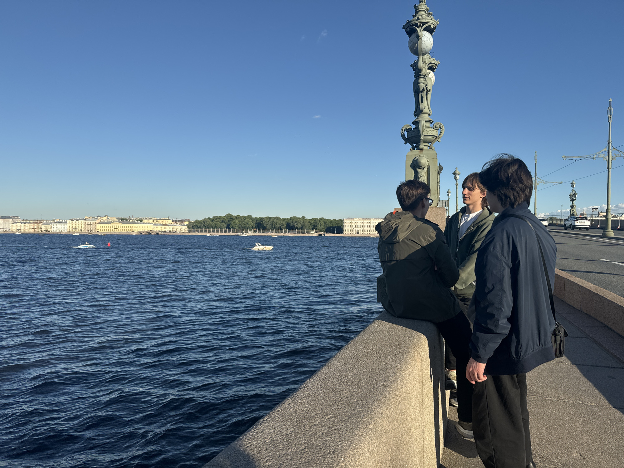 Three young men are standing on a bridge in Saint Petersburg, Russia, overlooking a wide body of water, likely a river or canal. The man on the left is sitting on a stone railing, facing away from the camera and towards the water. He is wearing a dark green jacket and dark pants. The man in the middle is standing and looking at the man on the left, wearing a light green jacket over a white t-shirt and dark pants. The man on the right is also standing and facing slightly away from the camera, wearing a dark blue jacket and black pants, with a black shoulder bag.
In the background, across the water, is a cityscape with buildings featuring classical architecture, likely part of the historical center of Saint Petersburg. A few boats are visible on the water. The sky is clear and blue, indicating a sunny day. A decorative lamppost, characteristic of Saint Petersburg's bridges, stands to the left of the group of men. The bridge appears to be a pedestrian walkway with a roadway for vehicles to the right.