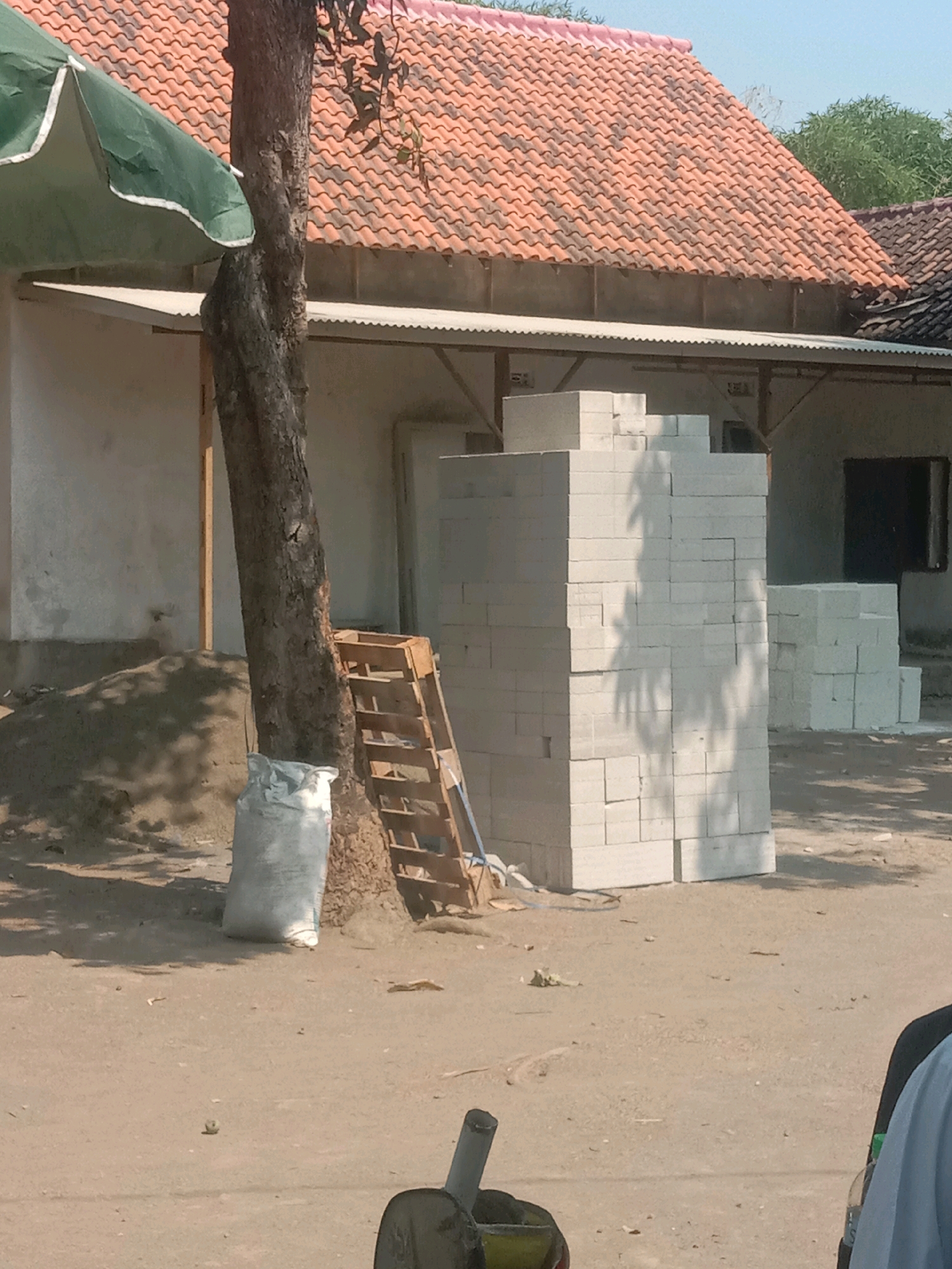 The image captures an outdoor scene in Ngempit, Indonesia, likely a construction site or material storage area. The focal point is a stack of white, rectangular building blocks. To the left of the blocks, a large tree trunk stands prominently, casting shadows on the ground. Adjacent to the tree, a white sack is visible, possibly containing building materials. A wooden pallet stands propped against the tree. Behind these elements, a building with a terracotta tile roof is partially visible, along with a section of a lighter-colored wall. The ground is predominantly dirt or sand, with scattered debris. In the foreground, a partial view of what appears to be a person's shoulder wearing a light blue garment is visible, suggesting human presence. The overall environment appears to be sunny and open.