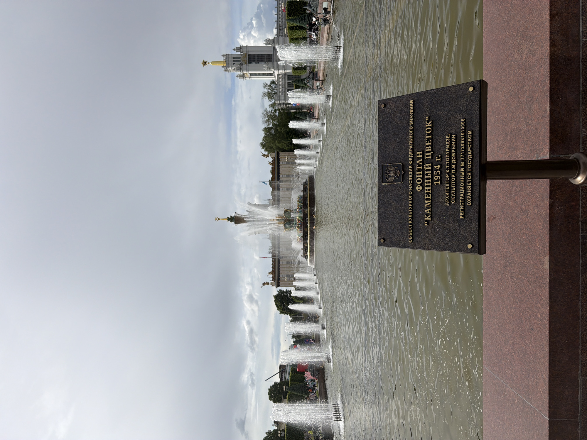 Here is a description of the image:
The image shows a fountain in Moscow, Russia. The "Kamennyy Tsvetok" ("Stone Flower") fountain, built in 1954, is shown with a plaque describing it as an object of cultural heritage under federal protection. The image shows the fountain with the water and jets spouting upwards, and in the background, there are buildings with gold stars on top. The image is rotated 90 degrees clockwise.