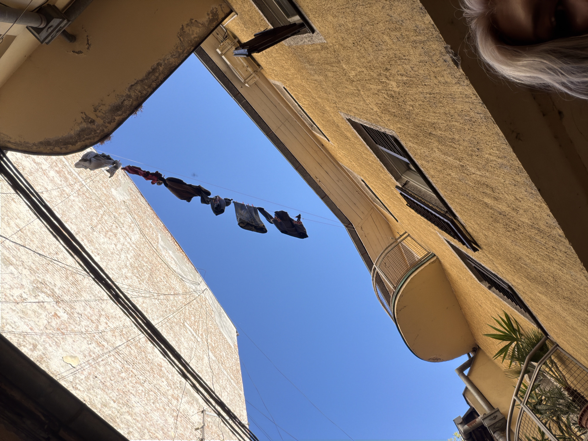 The image depicts a low-angle view in Belgrade, Serbia, looking up at buildings. Clothes are hanging on a line strung between the structures. The building on the left has a brick facade with electrical wires running across it. The building on the right is tan stucco and has a balcony with plants. A small portion of a person's head with blonde hair is visible in the upper right corner. The sky is clear and blue.