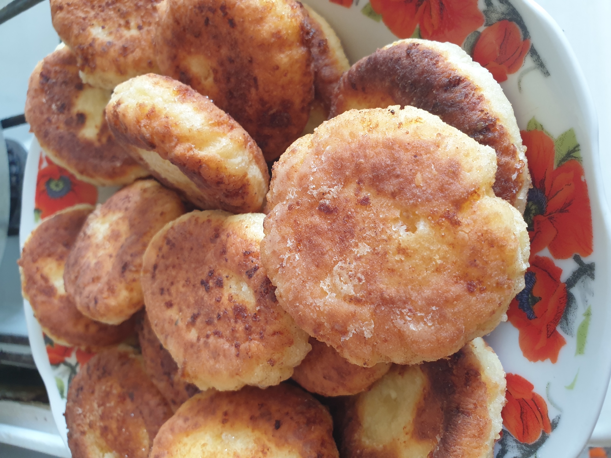 A close-up, overhead shot captures a stack of golden-brown, circular fried pastries on a decorative plate. The pastries, which appear to be syrniki (a type of Eastern European cheese pancake), have a slightly irregular, fluffy texture and are lightly dusted with sugar. The plate is white with a pattern of red poppies and green leaves. The background is out of focus, showing what appears to be a kitchen counter and possibly a stove. The lighting is natural, suggesting an indoor setting during the day. No people are visible.