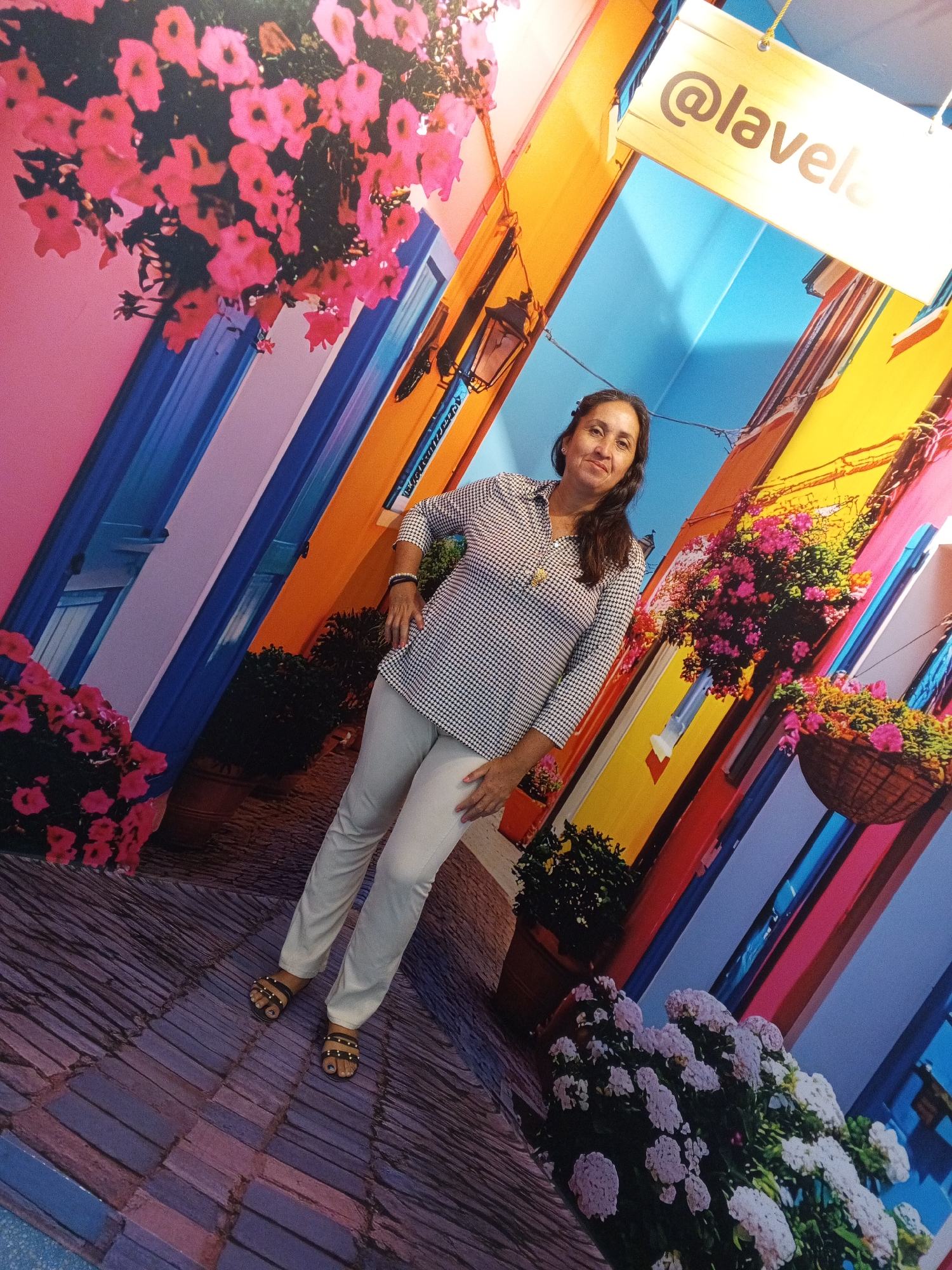 The image depicts a woman standing in a colorful, illusionistic setting designed to resemble a vibrant European street. The background features brightly painted buildings with exaggerated architectural details, such as windows and balconies adorned with flower boxes. The woman, wearing a black and white checkered blouse and white pants, stands with her left hand on her hip and her right arm at her side. She is looking towards the camera with a slight smile. In the foreground, the ground appears to be a cobblestone-like pattern, adding to the illusion of a street scene. Above her, a wooden sign with the text "@lavela" is visible, suggesting a location or brand name. The overall scene is characterized by its intense colors and playful design.
