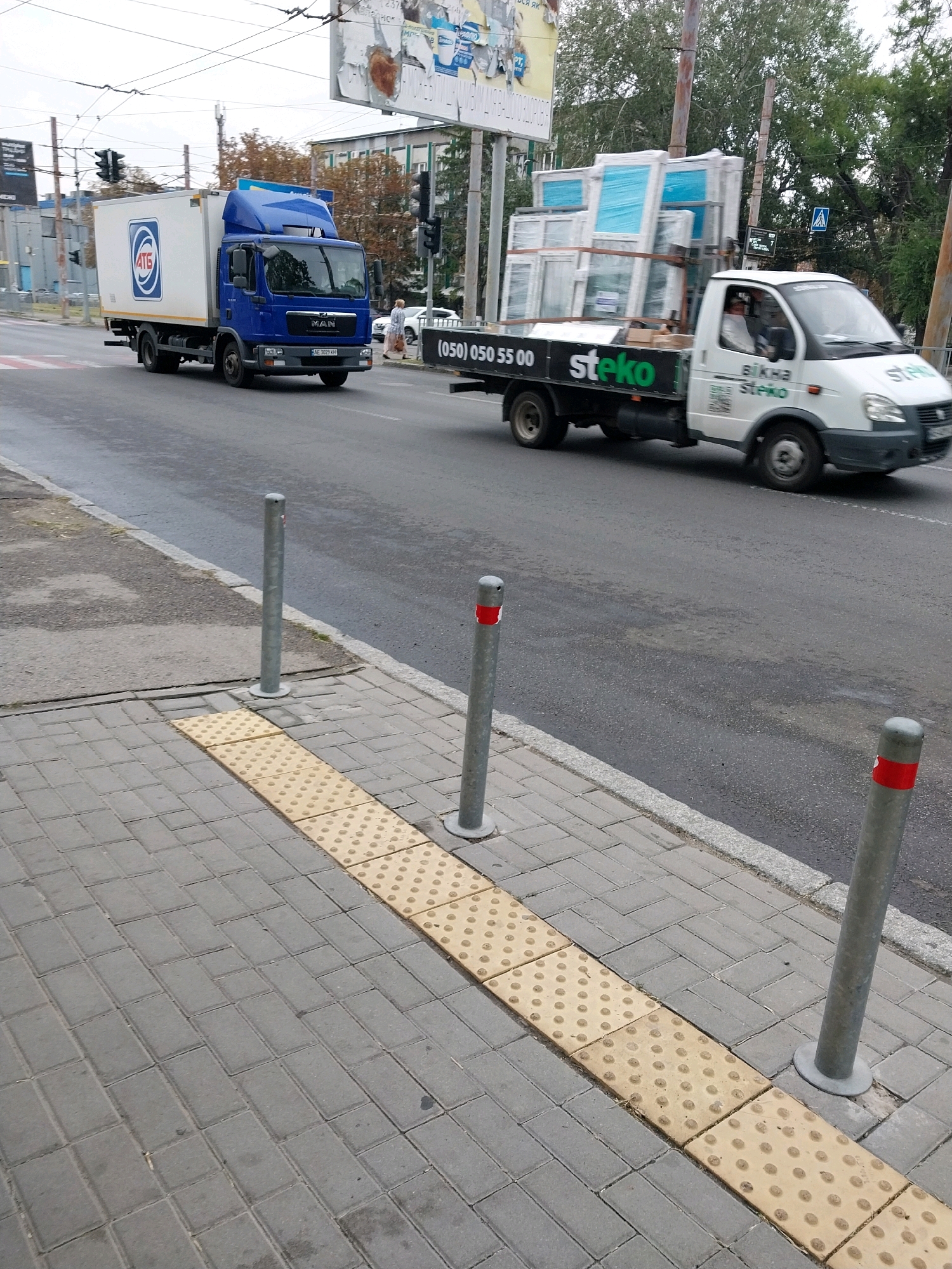 In an urban environment, two trucks are visible on a street. The first truck, a blue box truck with the logo "ATG" on its side, is moving left to right across the frame. The second truck, a white flatbed truck carrying several large windows, is also moving left to right, behind the first truck. The truck is branded with "Steko" on its side. The street appears to be wet, possibly from recent rain. The foreground shows a sidewalk with tactile paving, indicating a pedestrian walkway. Several metal bollards are present along the sidewalk, some with red reflective tape. The background reveals buildings, trees, and traffic lights, suggesting a city setting. The sky is overcast.