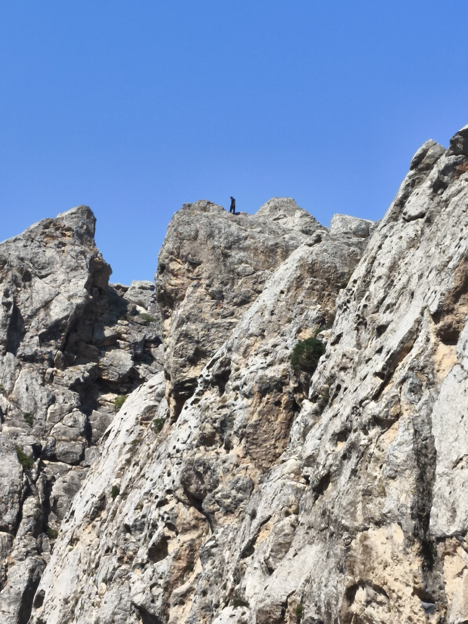 A lone individual stands atop a rocky cliff against a clear blue sky. The cliff face is rugged and textured, with visible strata and small patches of greenery. The person is silhouetted, making their features indistinguishable, but they appear to be facing away from the camera. The scene suggests a remote, natural setting. The location is identified as Qalaşıxı, Azerbaijan.