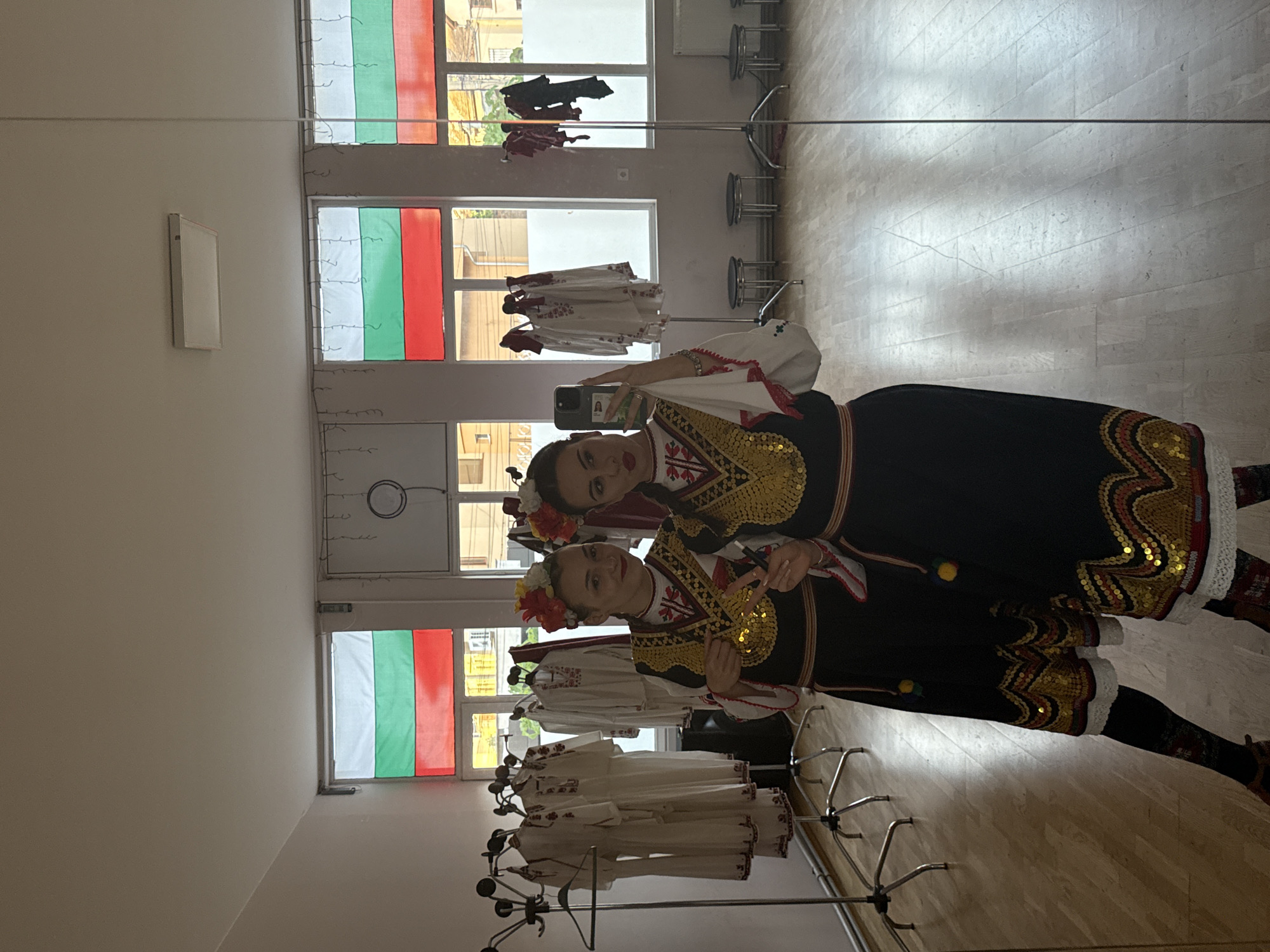 Two women are posing for a selfie in front of a mirror, in a room with a Bulgarian flag displayed in the windows. They are both wearing traditional Bulgarian folk costumes, adorned with intricate embroidery, gold accents, and floral headpieces. The women are positioned close together, suggesting a friendly or familial relationship. The woman in the background is holding the camera, taking the selfie, while the woman in the foreground is making a peace sign. Clothes hang on a clothing rack behind them. The reflection in the mirror shows the back of the room, revealing more windows, chairs, and items hanging on the rack. The location is inferred to be Mezdra, Bulgaria, based on the prominent display of the Bulgarian flag and the traditional attire of the women.