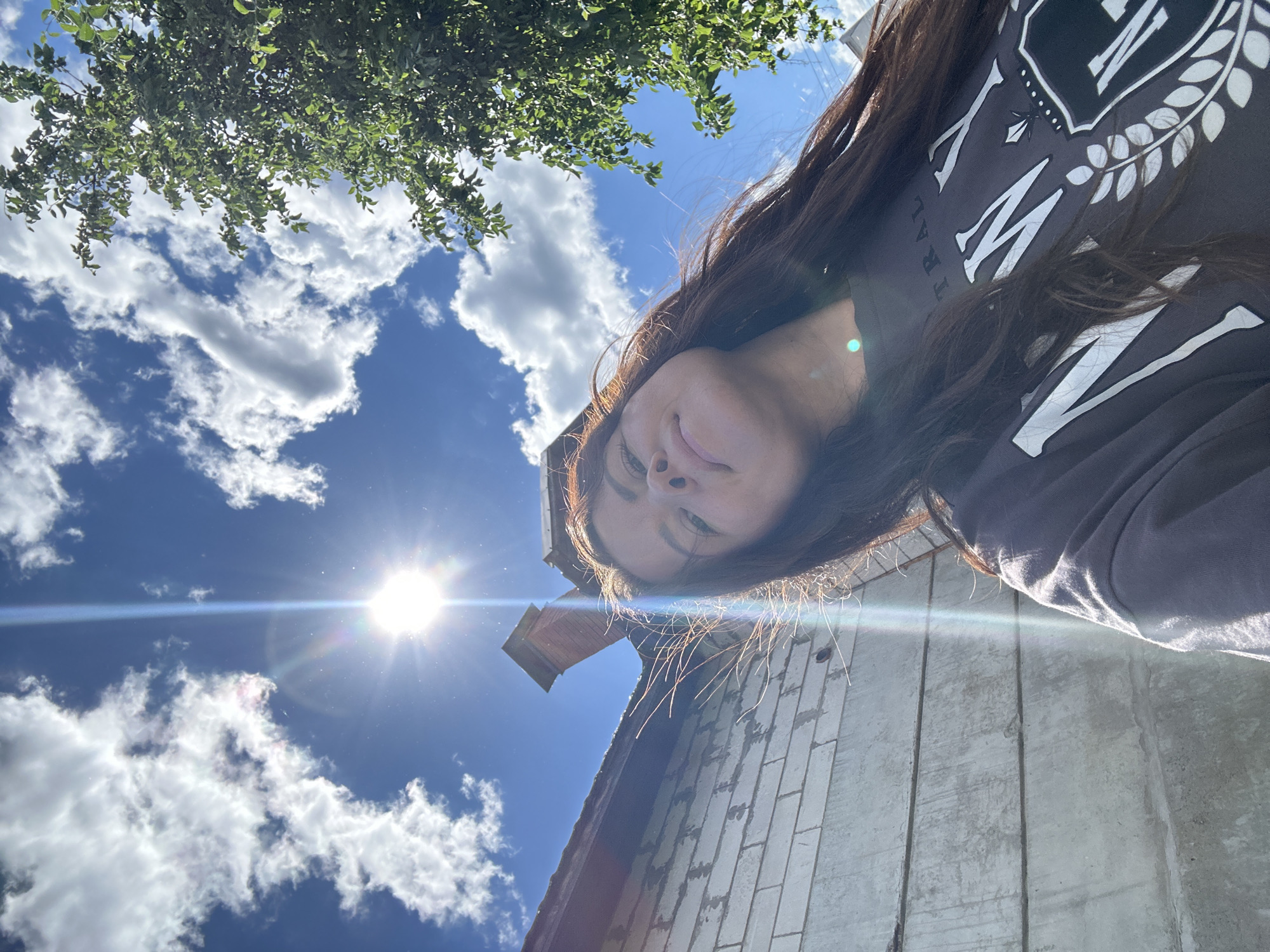 A young woman with long brown hair is pictured from a low angle, looking up towards the sun on a cloudy day. She is wearing a dark grey sweatshirt with white text and a crest. The background includes a brick chimney on a building, a light grey wall, and green foliage from a tree. The sun is shining brightly, causing lens flare. The scene appears to be outdoors in a residential or industrial area, possibly in the city of Chaikovsky, Russia, given the architecture and vegetation.