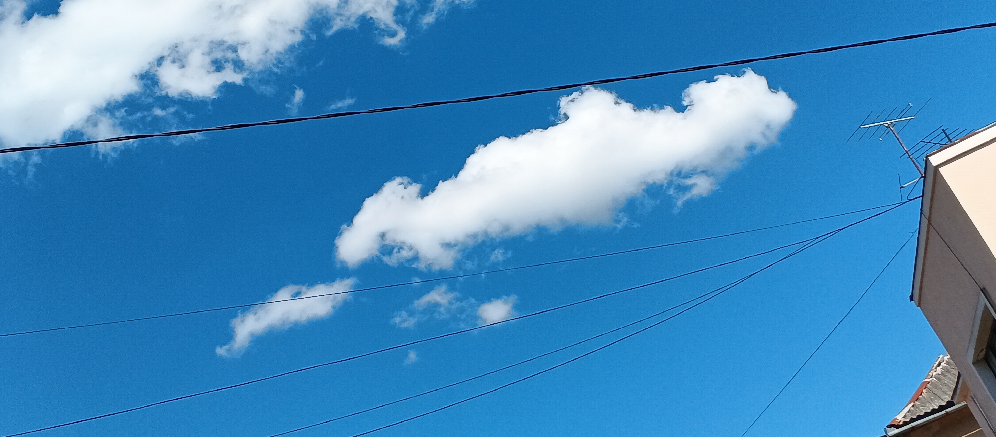 The image shows a bright blue sky with a few white clouds. Several dark electrical wires crisscross the sky at different angles. In the upper right corner, a portion of a building with a beige facade is visible, along with a television antenna mounted on its roof. The lower right shows a glimpse of a tiled roof. The scene appears to be outdoors in an urban or suburban setting. The location is identified as Mukachevo, Ukraine.