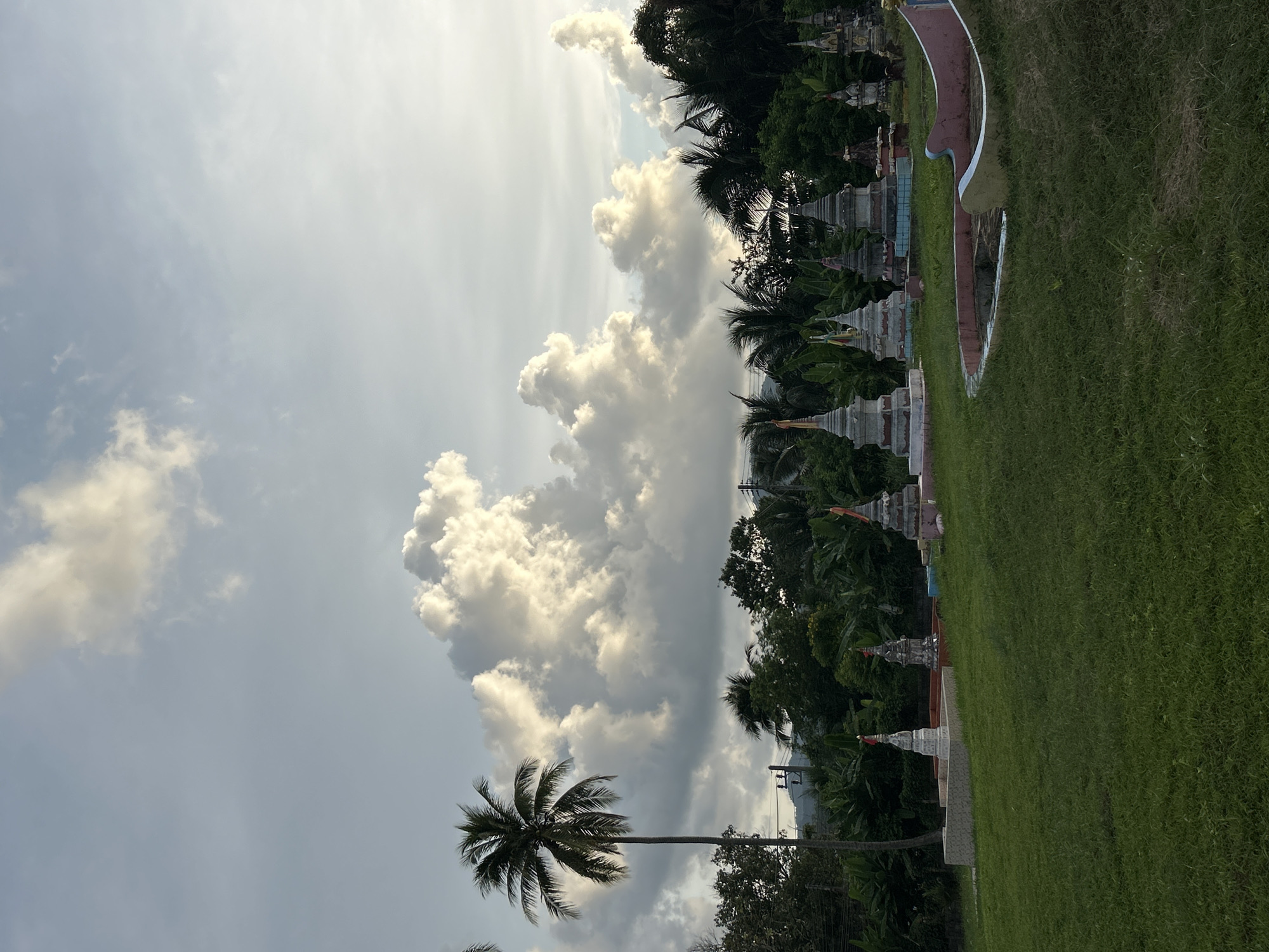 The image depicts a serene landscape scene, likely in Kathu, Thailand, under a sky filled with dramatic, illuminated clouds. In the foreground, a neatly mown green lawn stretches across the frame, with a curved, pink and light blue structure partially visible on the right. Scattered across the lawn are several small, ornate stupas, each topped with a colorful flag. A tall, slender coconut palm tree stands prominently on the left side of the image, its fronds silhouetted against the sky. Behind the lawn, a dense line of lush green trees and vegetation forms a backdrop, with glimpses of what appear to be structures and possibly hills in the distance. The overall impression is one of peacefulness and tropical beauty, captured during what appears to be either sunrise or sunset due to the lighting on the clouds.