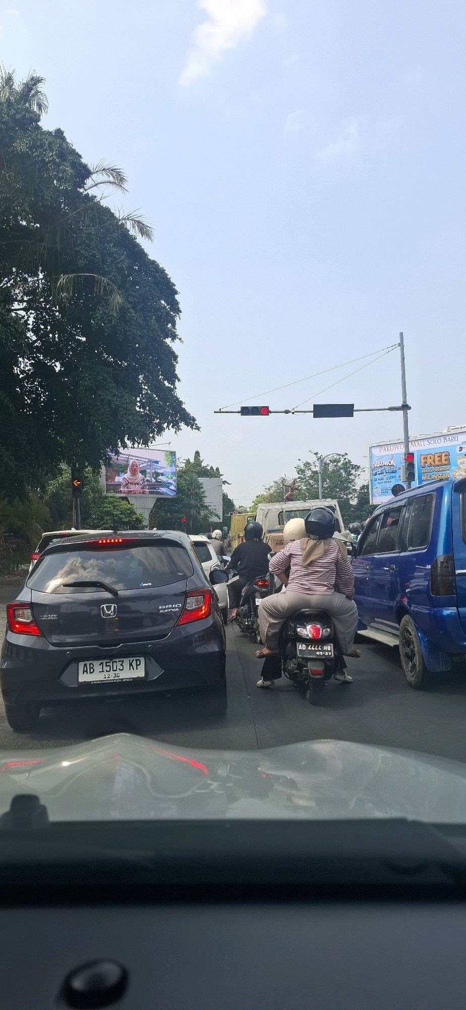 The image captures a traffic scene in Surakarta, Indonesia, likely on a sunny day given the bright sky with scattered clouds. The perspective is from inside a vehicle, looking forward through its windshield, which shows a distorted reflection of the surroundings.
In the foreground, the hood of the car the photographer is in is visible. Ahead, there is heavy traffic. A dark-colored hatchback, likely a Honda Brio Satya, is prominently in the left lane, displaying a license plate reading "AB 1503 KP". Beyond this car, other vehicles, including motorcycles and a blue minivan, are visible.
On the right side of the road, a blue minivan is parked or moving slowly. Several motorcycles are in traffic, with one carrying three people: a driver and two passengers wearing helmets and casual clothing. Another motorcycle behind it has a driver wearing a black helmet.
In the background, there are billboards and traffic lights. A large digital billboard displays an advertisement with a woman. Traffic lights are visible, with the one on the left showing red, and another further down the road also illuminated red. Trees line the side of the road, and utility poles with wires span across the scene. The overall impression is a typical urban traffic jam during daytime hours in Surakarta.