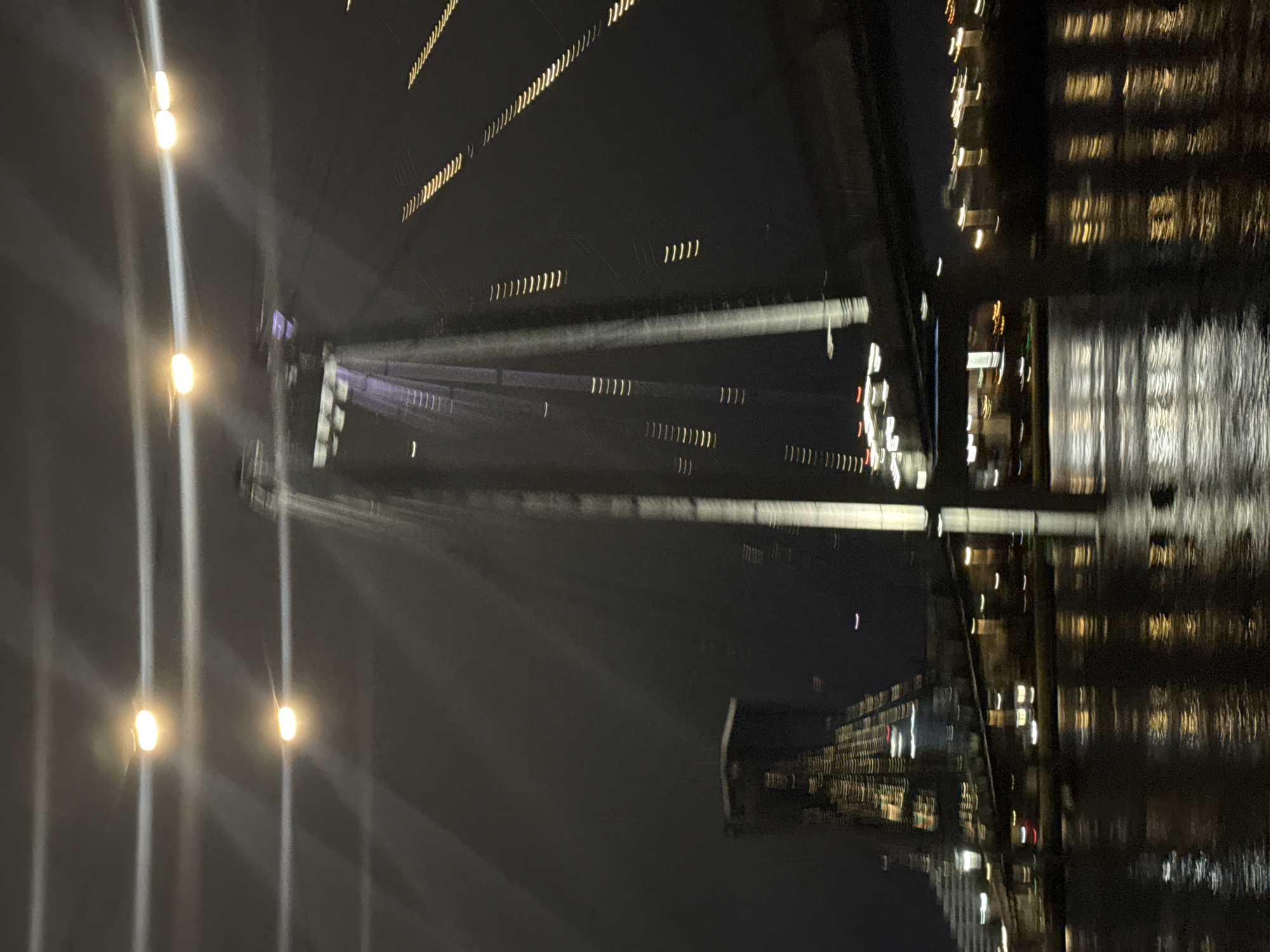 The image shows the Vinogradovsky Bridge in Krasnoyarsk, Russia, at night. The bridge's cables are illuminated, stretching from the supports to the roadway. Building lights reflect in the Yenisey River below. The buildings appear blurred due to camera movement.