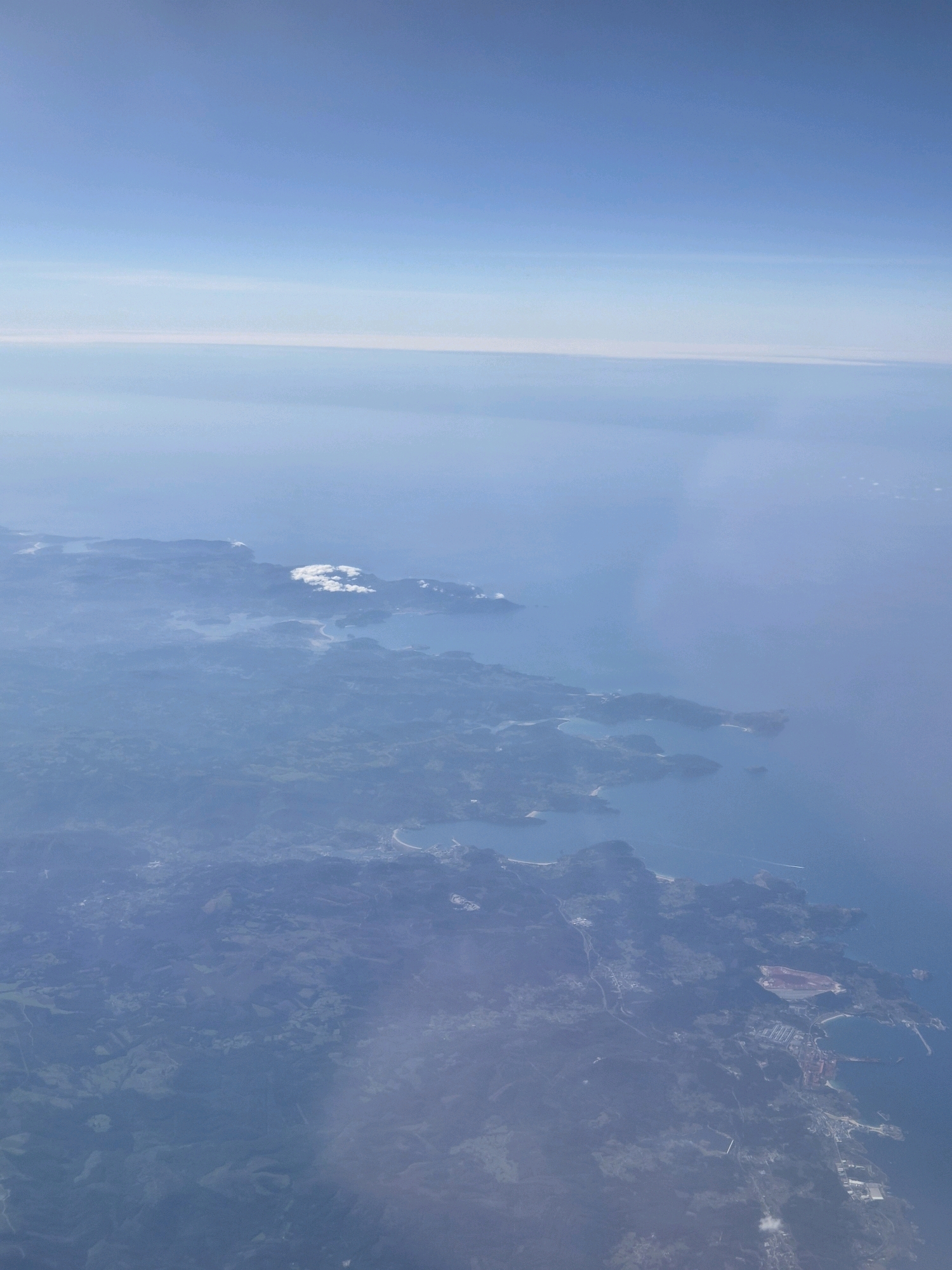 This is an aerial, high-angle, wide shot of a coastline on a hazy day. The sky is a gradient of light blue to a pale white horizon. The ocean is a deep blue and occupies the right side of the frame. The land is covered in lush green vegetation, with rugged coastlines, inlets, and small islands. White clouds are scattered over the land and ocean. Buildings are visible along the coastline, including some industrial facilities and what appears to be a town or village. A winding road is visible on the land. The scene is devoid of people and human activity. The image does not contain any identifiable landmarks or features that would allow for precise location identification in Foz, Spain.