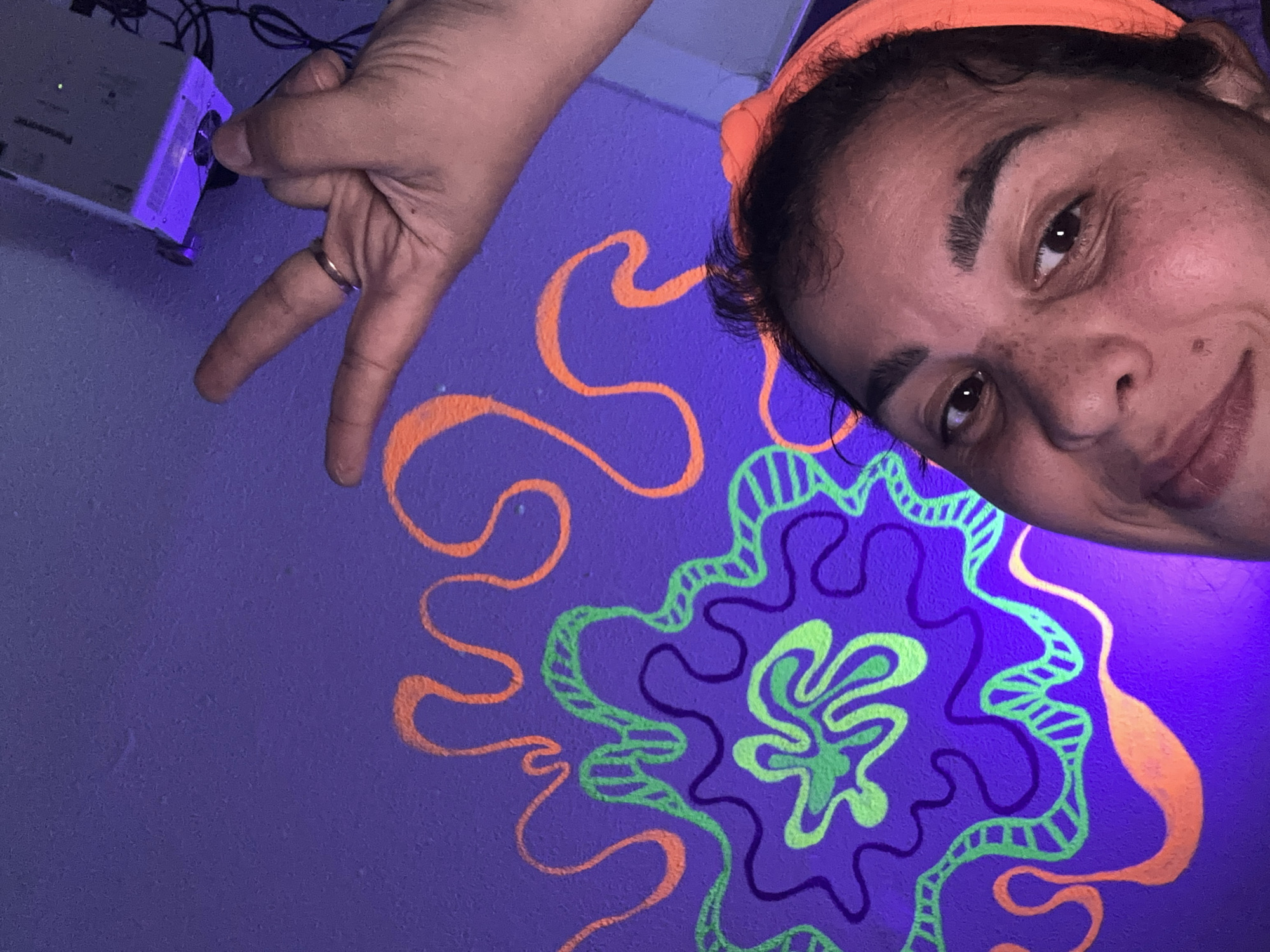 The media file depicts a close-up of a person's face and a hand against a purple wall adorned with neon orange and green artistic swirls. The person, with dark curly hair and light brown eyes, is wearing an orange headband and appears to be smiling or smirking. Their face is tilted, and they are looking towards the camera. Freckles are visible on their cheeks and nose. The hand, with a gold ring on one finger, is positioned in the frame, making a peace sign gesture. To the left, a portion of a white electronic device, possibly a projector, is visible. The overall scene is illuminated by what appears to be a black light, causing the neon colors of the wall art to glow vibrantly. The purple background of the wall suggests an indoor setting, possibly a room decorated for a party or artistic display. The mood appears playful and candid. No visible text is present in the media file.