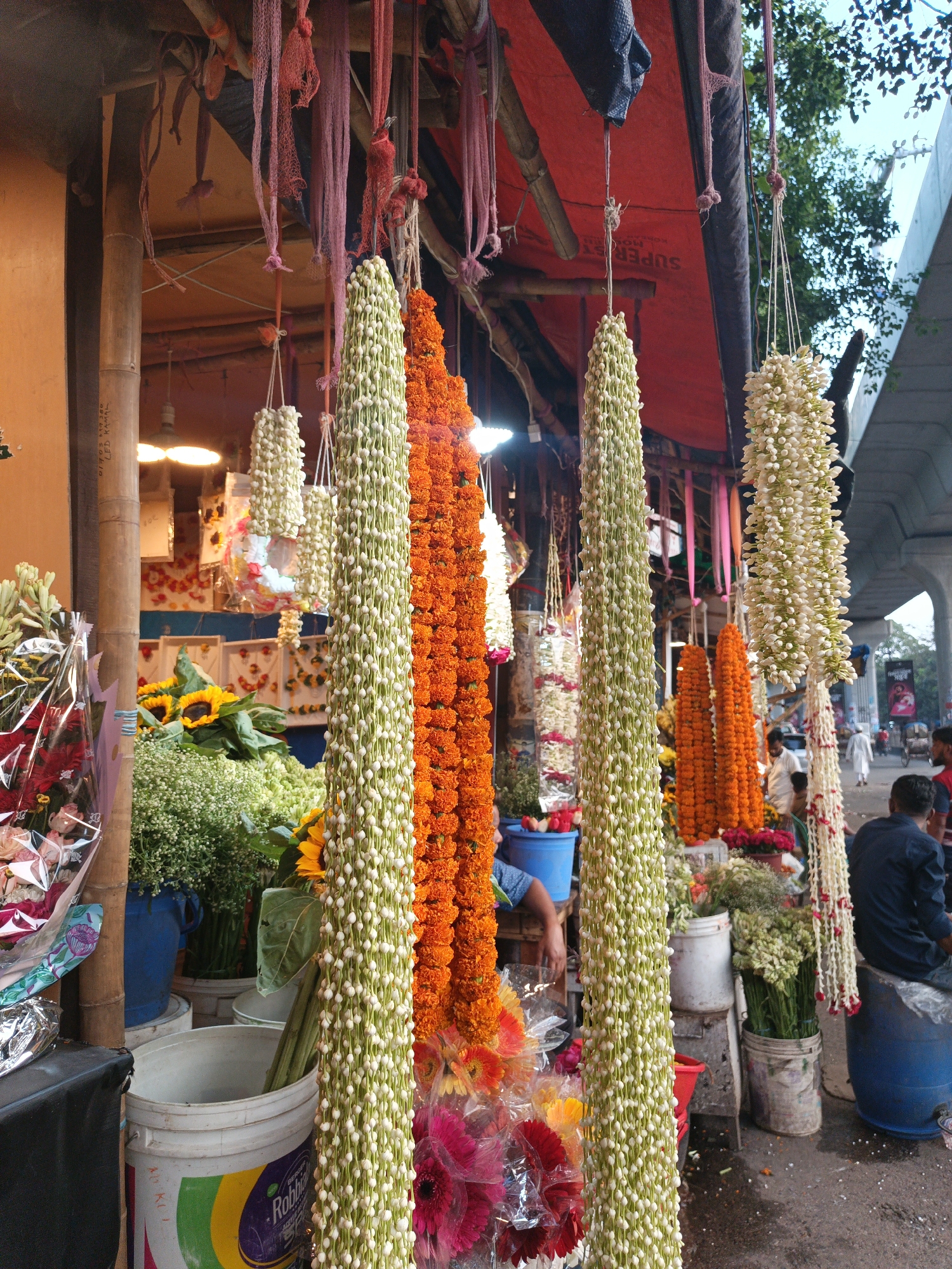 This eye-level outdoor shot shows a flower shop in Dhaka, Bangladesh. Garlands of flowers, white and green and orange, hang from a red-roofed storefront that has pink and red netting decoratively strung along the top. There are vases and buckets of cut flowers, including sunflowers, roses, and greenery. A man in a blue shirt sits on a blue barrel on the right side of the shot. Other people can be seen in the background, including a man in a white shirt and a woman in a pink sari. A highway overpass looms in the background on the right side of the image.