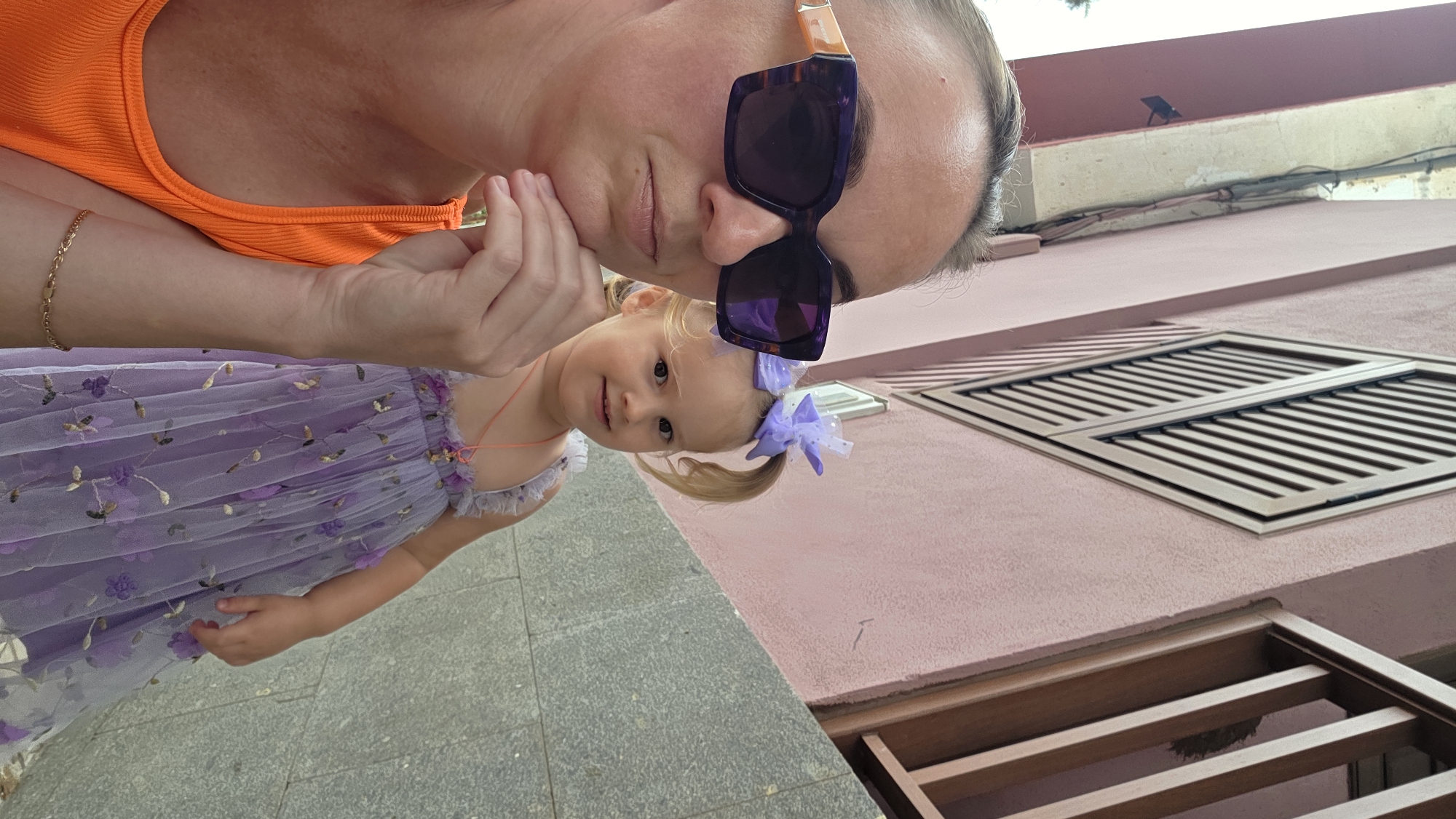 In the city of Guía de Isora, Spain, a medium shot captures a woman and a young child outdoors. The woman, positioned on the left, wears an orange sleeveless top and large purple sunglasses. Her right hand is raised to her chin, and she has a neutral expression. To her right, the young child is dressed in a lilac tiered dress adorned with floral embroidery. The child has blonde hair styled in two pigtails, each secured with a purple bow, and wears a pink pendant necklace. The background features a building with a pink facade, featuring a diamond-shaped window shutter and a wooden railing. A grey stone wall runs along the lower portion of the image. The lighting suggests it is daytime, with diffused natural light.