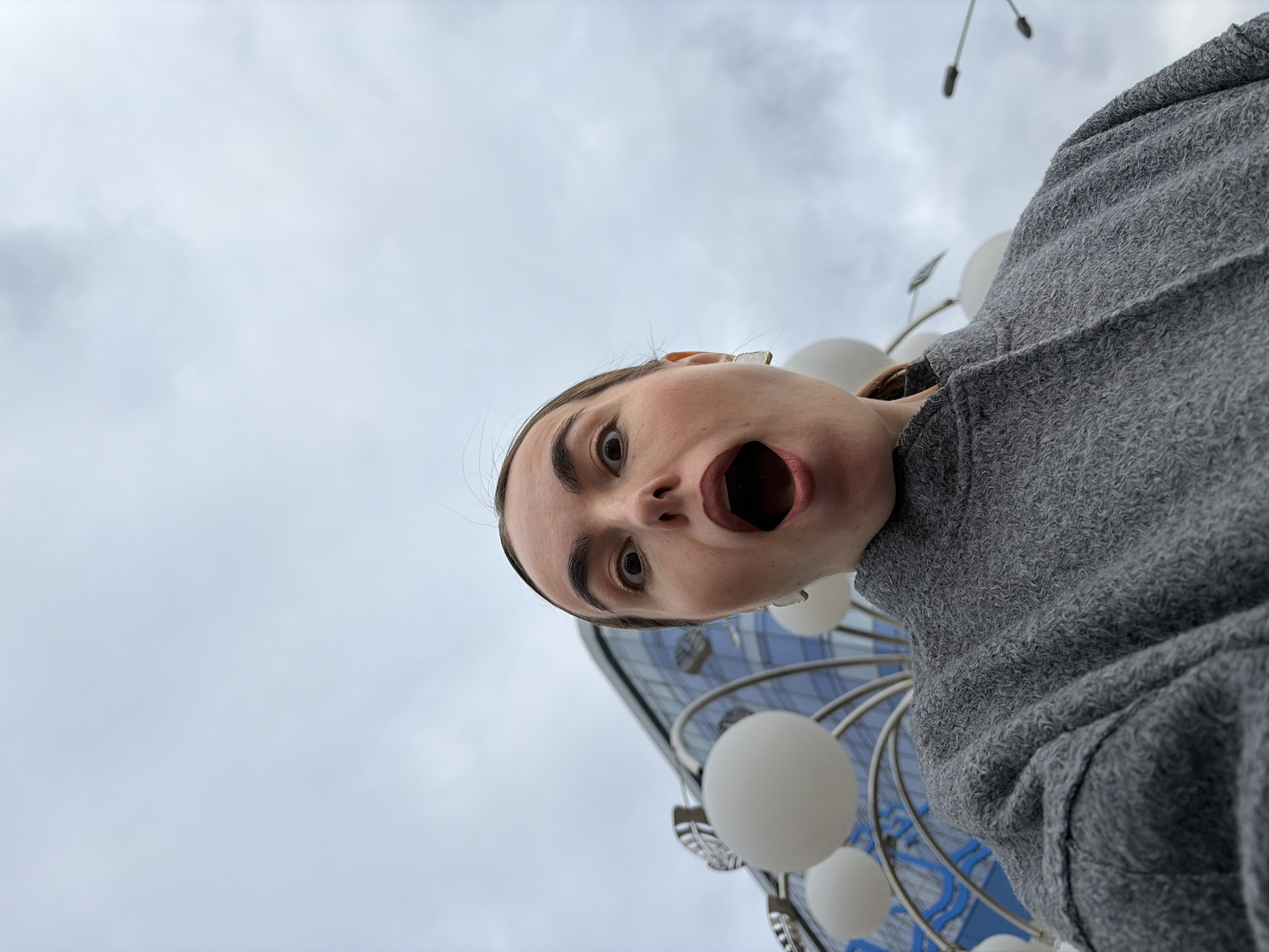 A person with an open mouth, likely in a surprised expression, is captured in a low-angle, slightly tilted close-up shot. The individual has light skin, dark eyebrows, and dark hair pulled back. They are wearing a gray textured garment.
In the background, partially visible, is a modern building with a glass facade and a curved design. In front of the building are decorative light fixtures featuring round, white lamps or globes mounted on metal structures. The sky is overcast with gray and white clouds.
Based on the architecture of the building and the style of the light fixtures, the location is likely the Central Square in Izhevsk, Russia.
