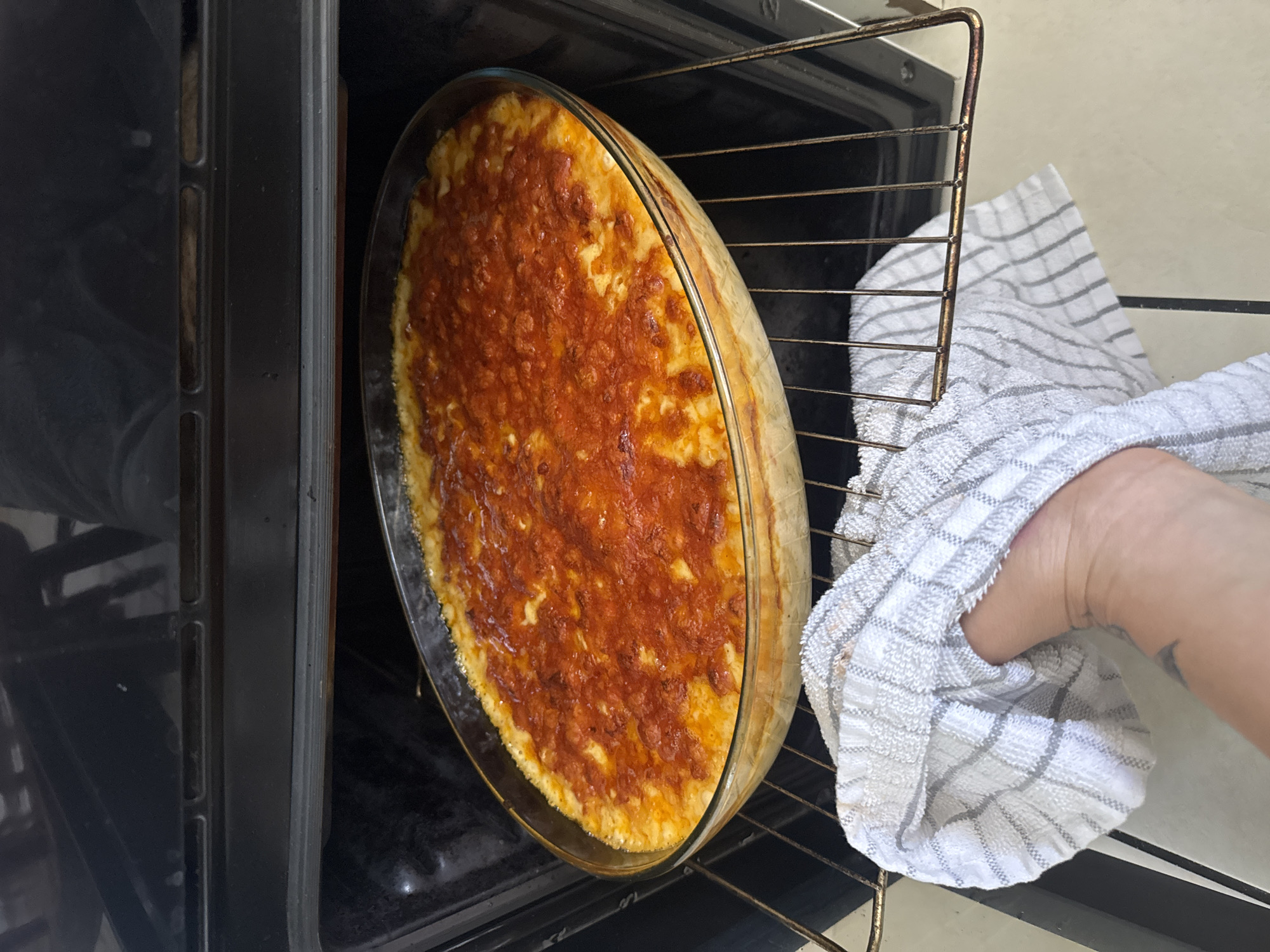A glass baking dish filled with a golden-brown, bubbly casserole topped with melted cheese and possibly a red sauce is positioned on the middle rack of a dark-colored oven. A checkered white and gray dish towel is being used by a person to partially support the dish, which is positioned for removal or insertion. The oven interior is visible, with metal racks and a dark frame. The environment appears to be a kitchen. The location is not specified in Armagh, United Kingdom.