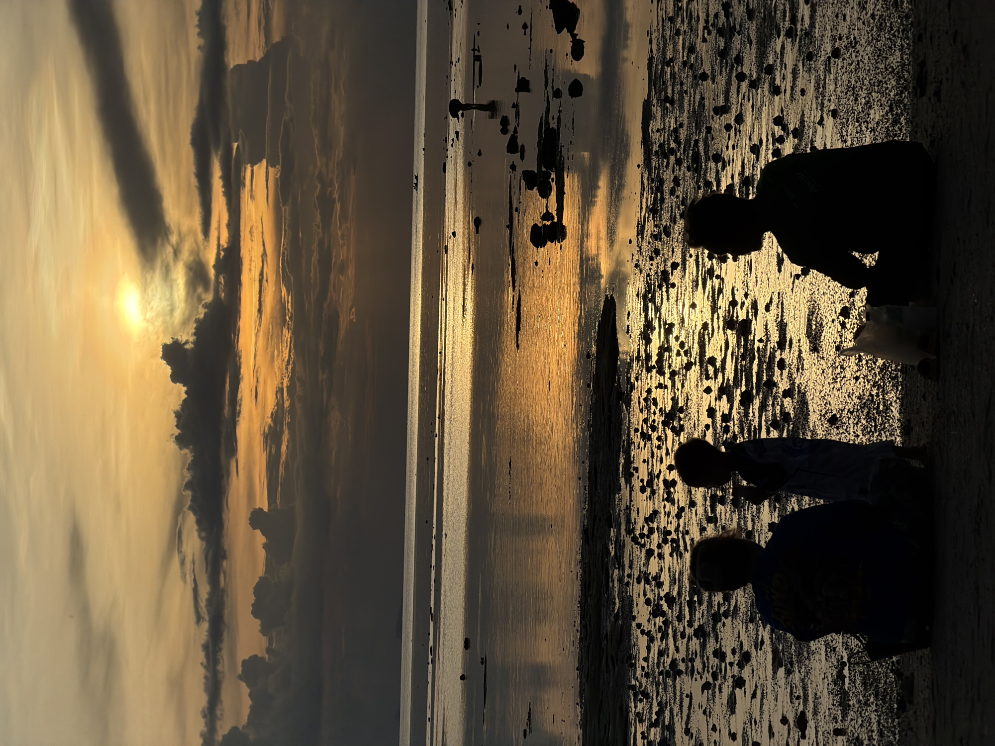 The image depicts a sunset scene at the beach in Baan Plai Laem, Thailand. In the foreground, silhouetted against the vibrant sky, are three people and a small dog sitting on the sand. Two adults are seated, and a child stands between them, looking towards the ocean. The sand is strewn with numerous small, dark rocks and pebbles. The shallow water of the sea stretches out to the horizon, reflecting the warm hues of the sunset. In the distance, the sun is partially obscured by dramatic, dark clouds, casting a golden glow across the sky and the water. There are small, dark objects visible in the water, possibly rocks or debris, and a faint silhouette of a boat on the horizon. The overall atmosphere is one of peaceful contemplation as the day ends.
