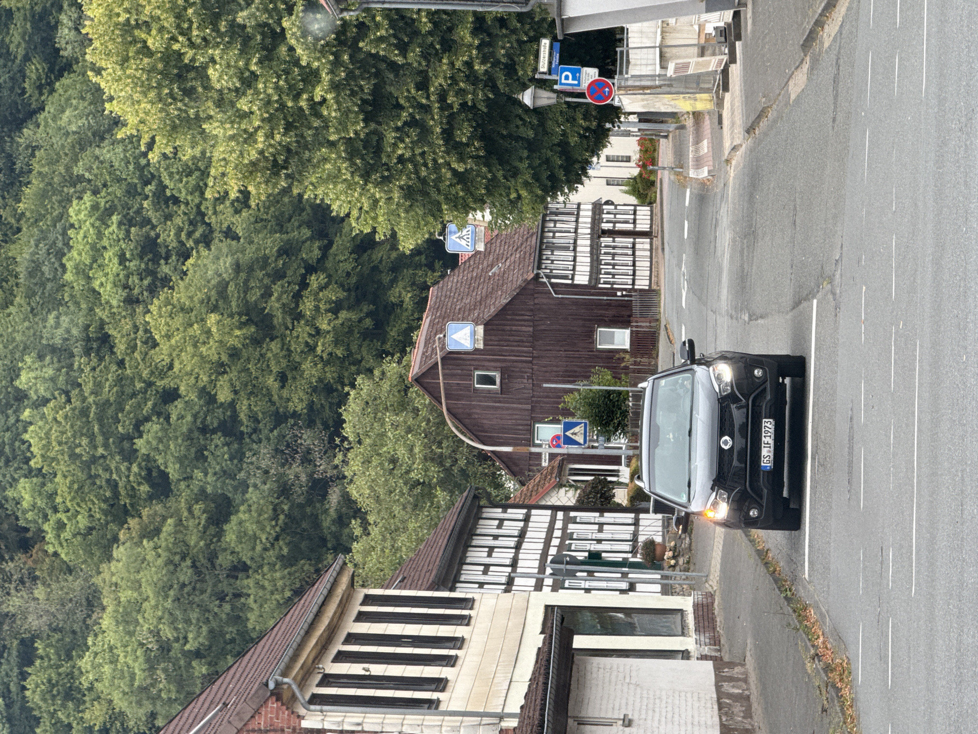 The image depicts a street scene in the city of Bad Lauterberg im Harz, Germany. A black SUV is driving towards the camera on a paved road with white lane markings. The car has its headlights on and its license plate reads "GS IF 1973". On the left side of the road, there are traditional half-timbered houses with white facades and dark wooden beams. To the right of the road, there is a large, dark brown wooden barn-like building and a tall, leafy tree. Further down the street, a blue parking sign is visible, along with a red and blue circular sign with an arrow. The background is dominated by a dense, green forest covering a hillside. The environment appears to be a somewhat rural or historic part of a town.