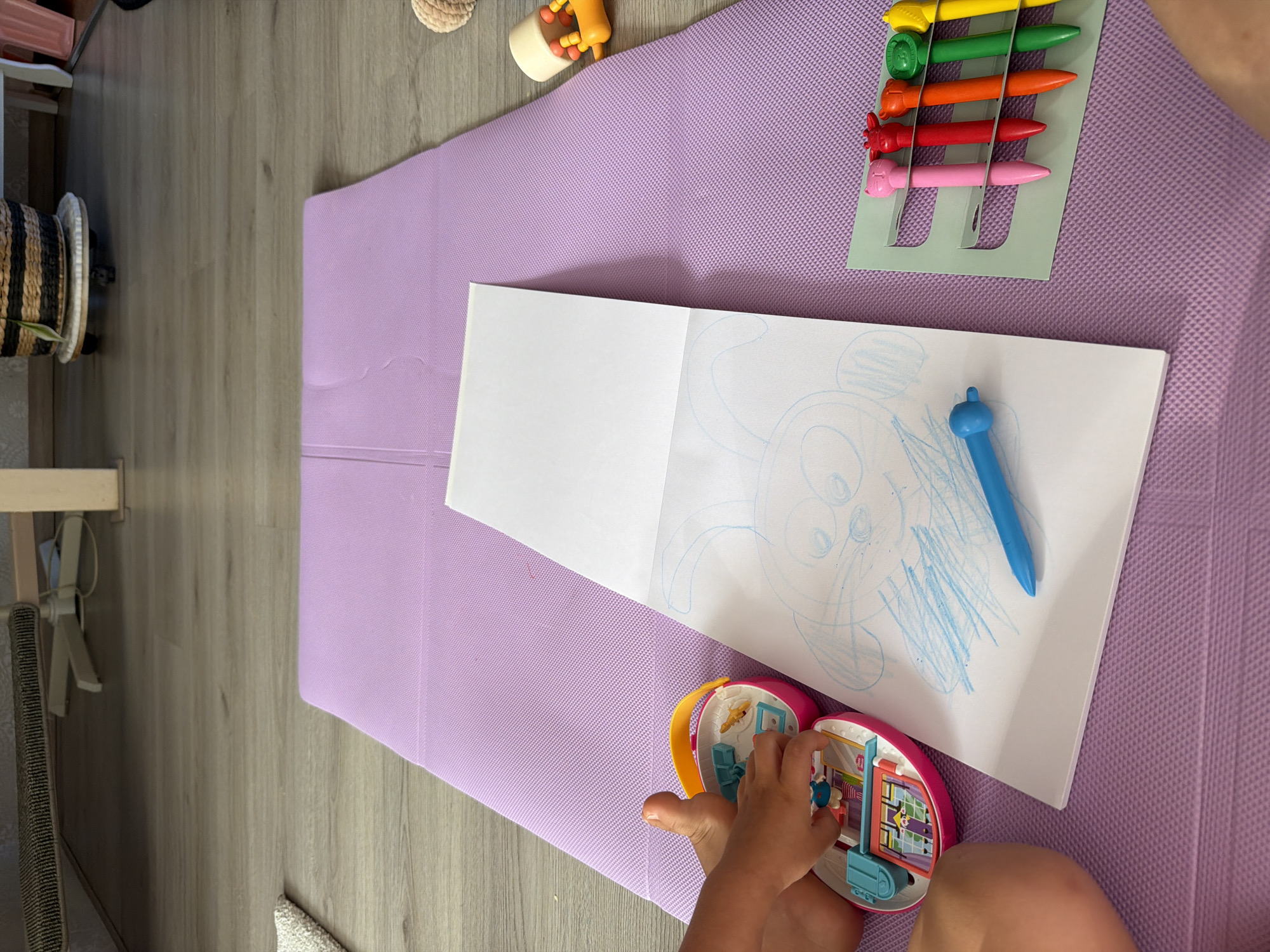 A young child's left leg and hand are visible on the bottom left of the frame, positioned on a purple mat. The child is playing with a pink toy with a yellow handle, which appears to be a miniature dollhouse. Next to the toy is a white sketchbook with a blue crayon drawing of a bunny. A blue crayon rests on the drawing. To the right of the sketchbook, a green holder contains five colorful pens with animal-shaped tops: pink cat, red deer, orange fox, green bear, and yellow lion. A yellow toy with orange beads is visible on the purple mat to the right of the pens. The background shows a wooden floor and furniture, including a woven basket on a wheeled stand. The scene appears to be indoors, likely in a home in Melick, Netherlands.