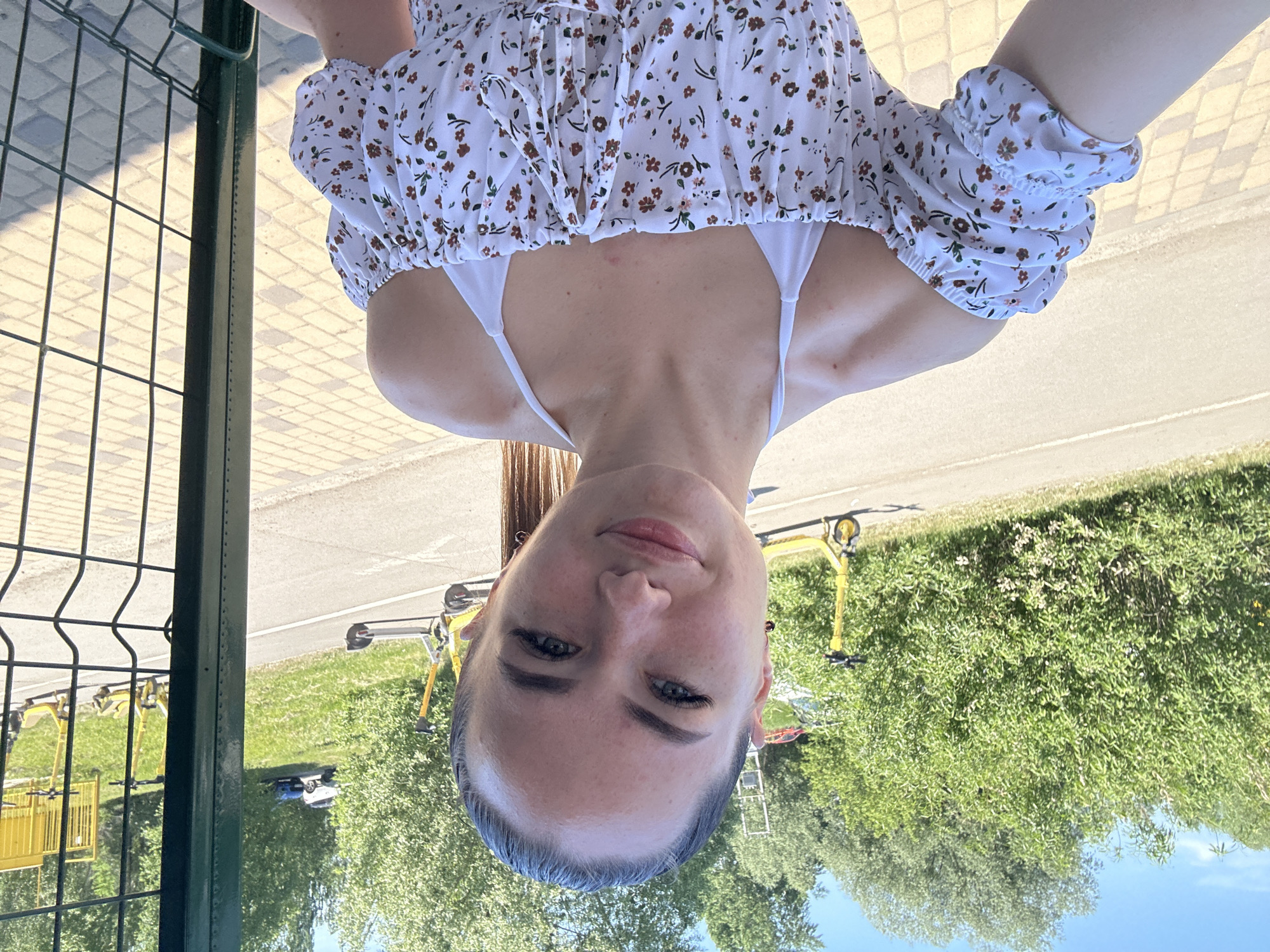 A young adult female is captured in a selfie on a bright, sunny day, standing outdoors in what appears to be a park or recreational area. Her dark brown hair is pulled back into a high ponytail, revealing a clear forehead with some natural flyaways. Her face is lightly freckled across the nose and cheeks, with blue-grey eyes accentuated by long eyelashes, and arched eyebrows. She wears small dark stud earrings and has a slight, closed-lip smile. She is dressed in a white off-the-shoulder blouse with short, puffy sleeves, patterned with small brown, orange, and pink flowers and green leaves. Beneath the blouse, a white halter-neck top is visible. Some faint redness or blemishes are present on her upper chest and shoulders.
The background features a paved asphalt path or road stretching horizontally, bordered by dense green bushes and taller trees under a clear blue sky. Several bright yellow electric rental scooters are distributed across the scene: one is prominently visible on the path to the left, another partially obscured behind the woman's head, and multiple others are lined up on the right side of the path, behind a dark green metal mesh fence. In the distant background, on a grassy incline, a small white utility vehicle and a blue vehicle are partially visible, along with a metal ladder. The woman is positioned to the left of a sturdy dark green metal fence post and mesh fence, which occupies the right portion of the frame. The immediate foreground where she stands is paved with yellow-tan square paving stones. Strong sunlight creates distinct shadows, indicating clear weather.