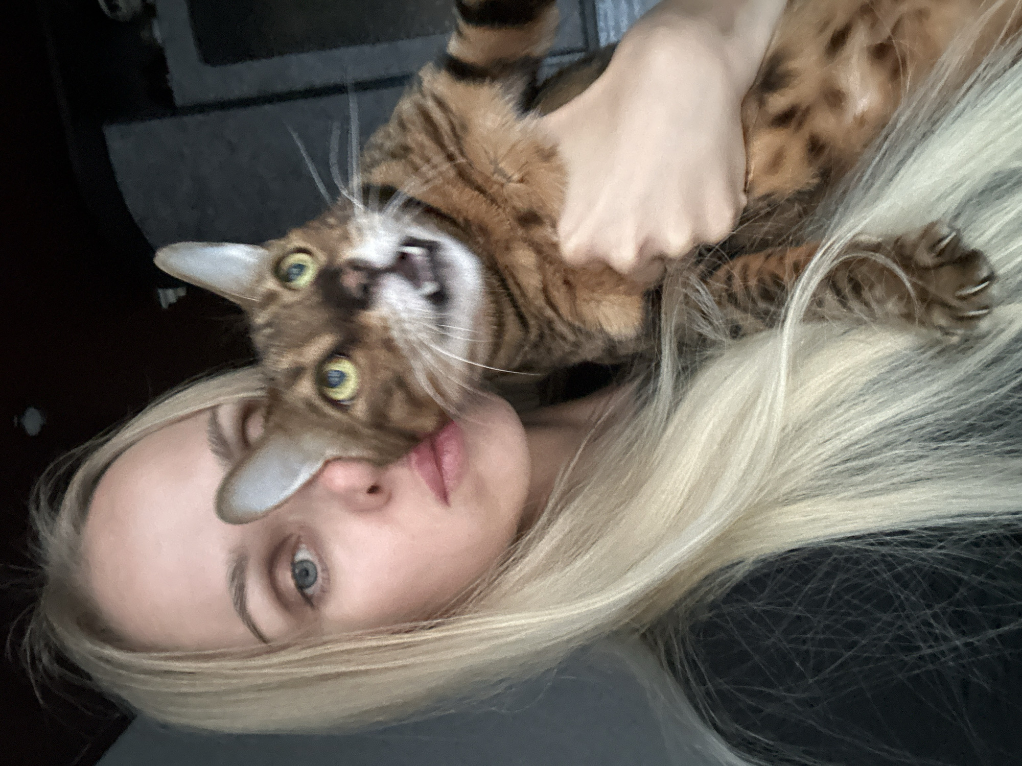A woman with long blonde hair, wearing a black shirt, is holding a Bengal cat in her arms. The cat is looking directly at the camera with its mouth open, showing its teeth. The background is dark and out of focus, with some shelving visible. The image appears to be a selfie taken indoors. The specific location in the city of Energetik, Russia, cannot be determined from the image alone.