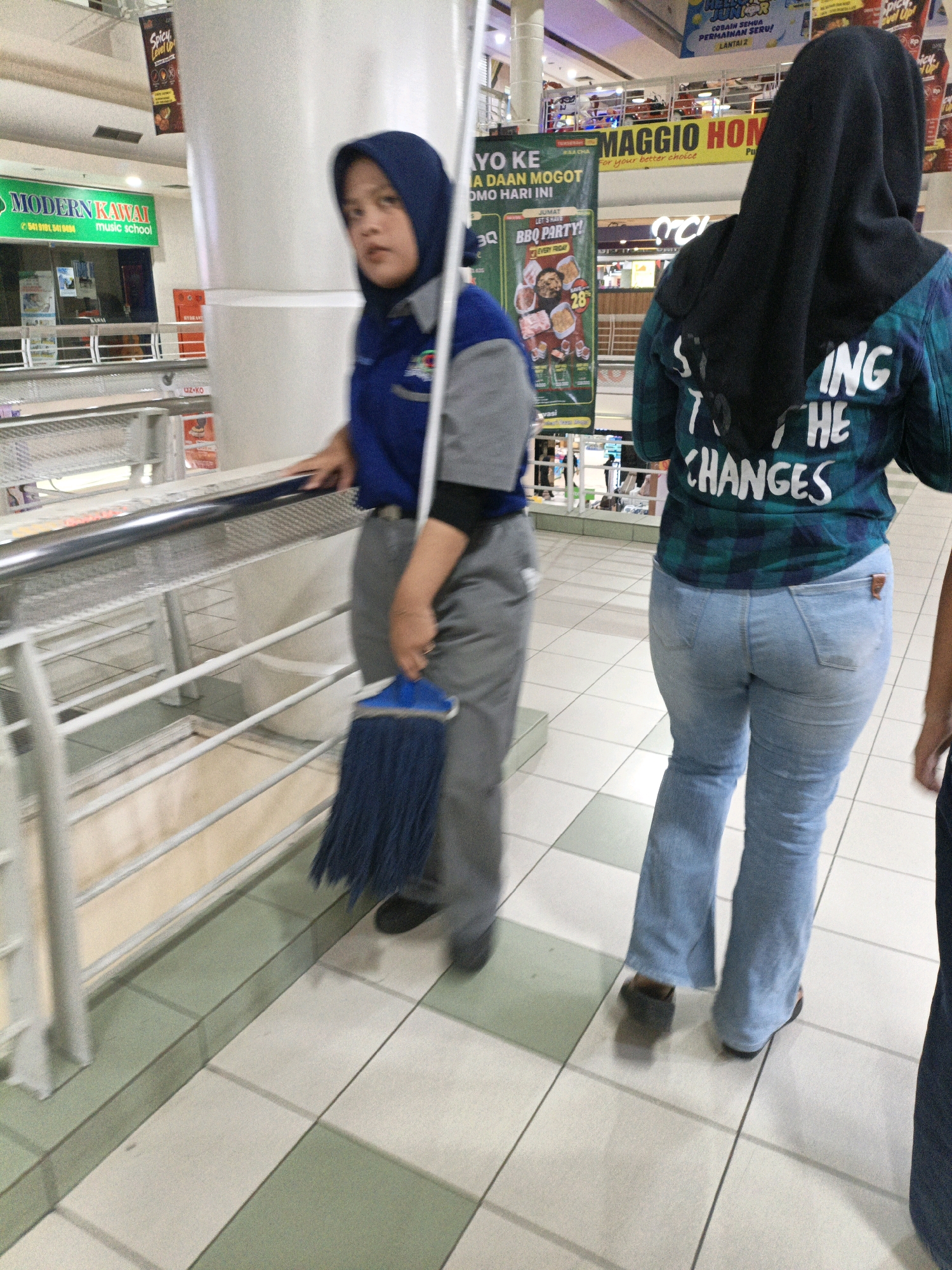 The image depicts an indoor environment, likely a shopping mall, in West Jakarta, Indonesia. Two individuals are visible. In the foreground and to the left, a person wearing a blue hijab and a blue and gray uniform is holding a blue mop. They are standing on a tiled floor with green and beige squares and leaning on a metal railing. To the right, another person is seen from behind, wearing a black hijab and a blue and green plaid shirt with white text that reads "START TO THE CHANGES". This person is wearing light blue denim jeans and dark footwear. In the background, there are advertisements and signs, including one that says "MODERN KAWAI MUSIC SCHOOL" and another with "MAGGIO HOME". The overall lighting suggests an artificial indoor setting.