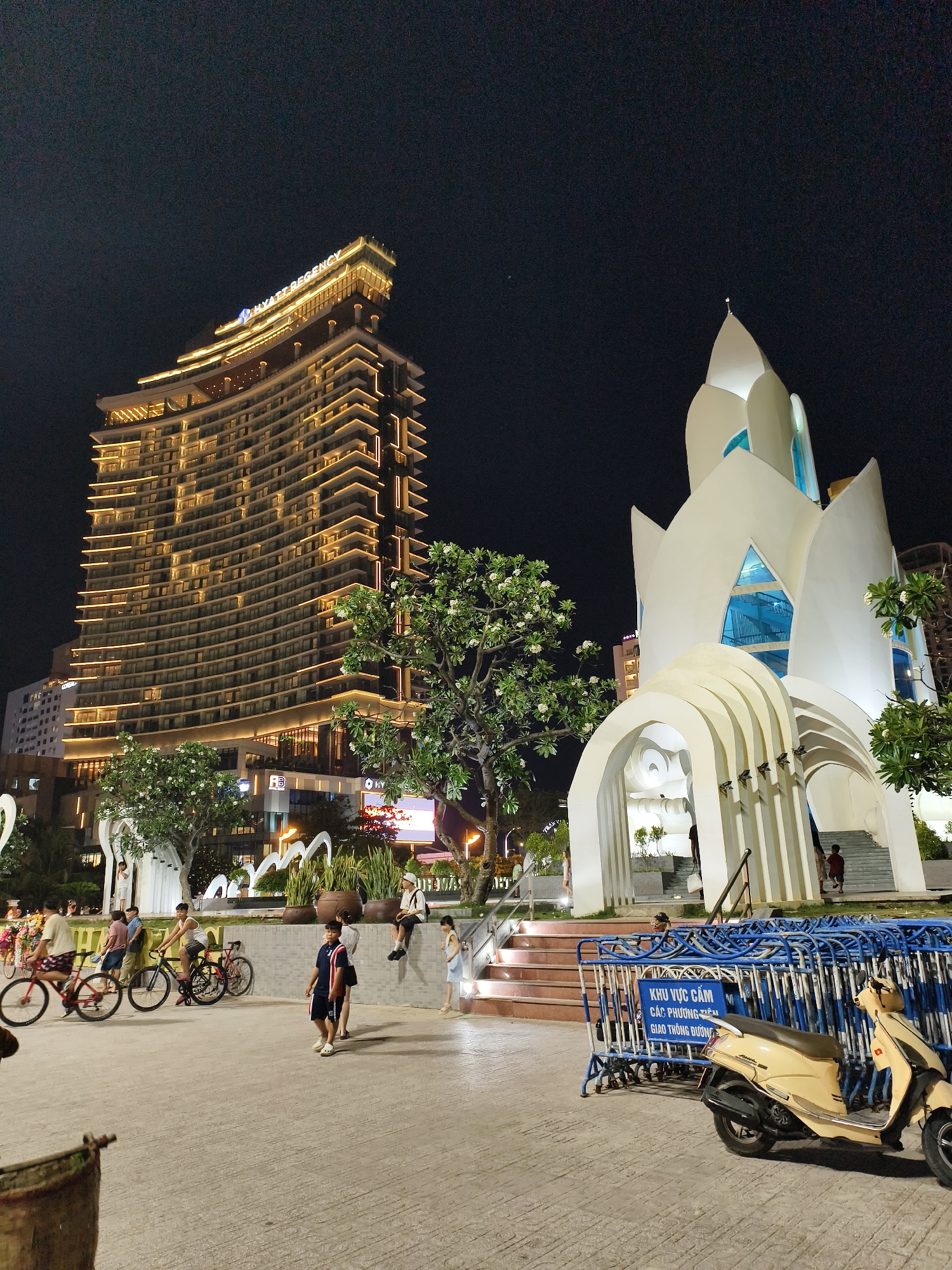 At night, a view of a plaza shows a building, a church, people on bicycles, pedestrians, a scooter, and temporary fencing. The large, modern, high-rise Hyatt Regency Hotel is on the left, brightly illuminated with vertical and horizontal bands of light emphasizing the building’s contours. A white church with a pointed spire and petal-like structures forming the upper portion is on the right. The church has open arches and blue glass accents. In front of the church, a temporary blue metal fence surrounds an area with a yellow scooter parked nearby. A sign in Vietnamese is attached to the fencing. People are walking and riding bicycles in the plaza. Some are on bicycles. A few sit on a raised ledge. Trees are interspersed between the buildings and people. The ground of the plaza is paved with concrete slabs. The scene is located in Nha Trang Ward, Vietnam.