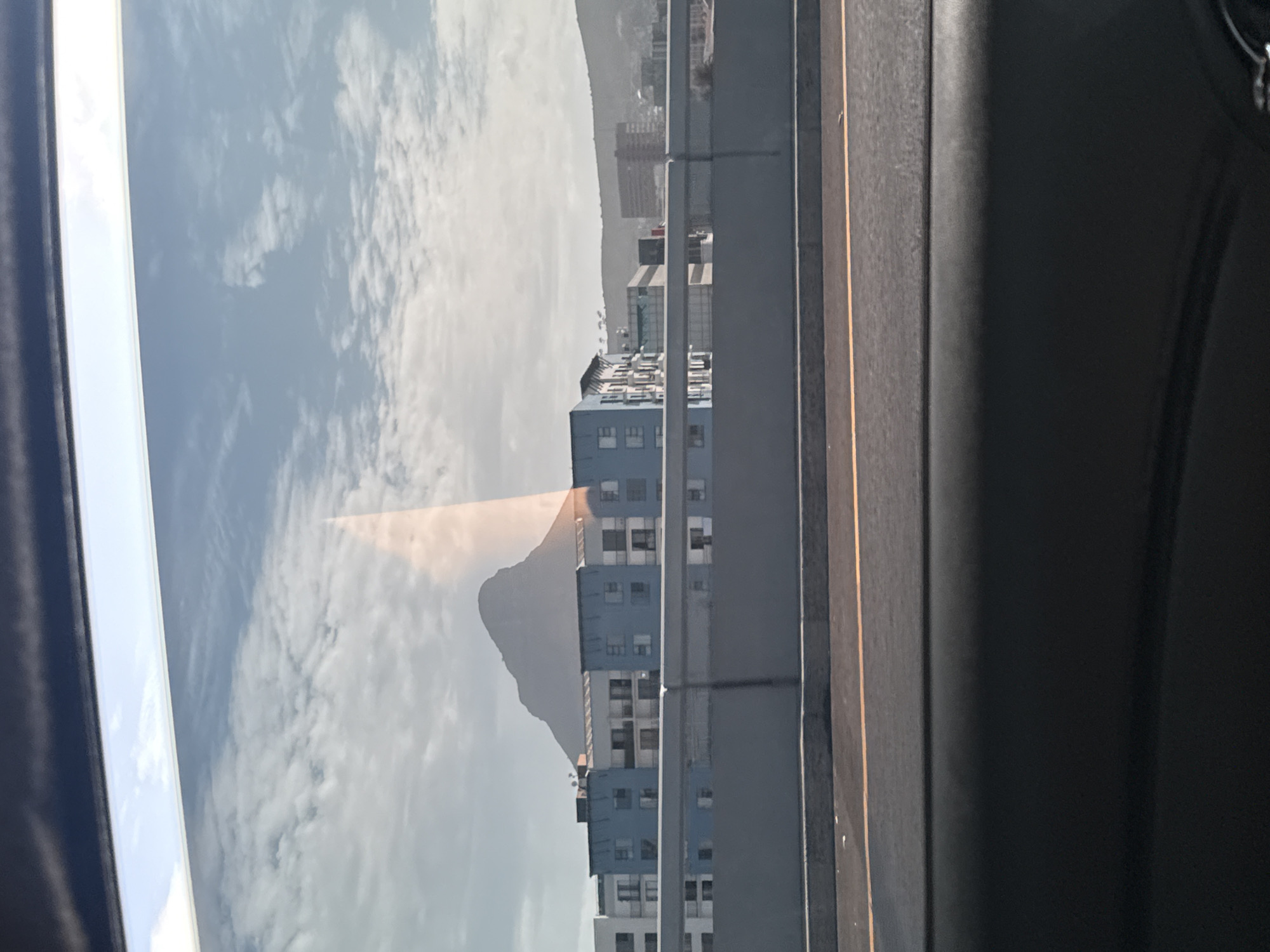 The image shows a view from inside a vehicle, likely on a highway or elevated road. Through the window, there's a reflection of a mountain, possibly Table Mountain, combined with the view of a building with multiple floors and windows. The building seems to be located near the highway, with a barrier or wall running along the side. The sky is visible, with clouds suggesting a bright day. The interior of the vehicle is dark, with portions of the dashboard and possibly the side of the car visible. The location is likely Cape Town, South Africa, considering the potential presence of Table Mountain.