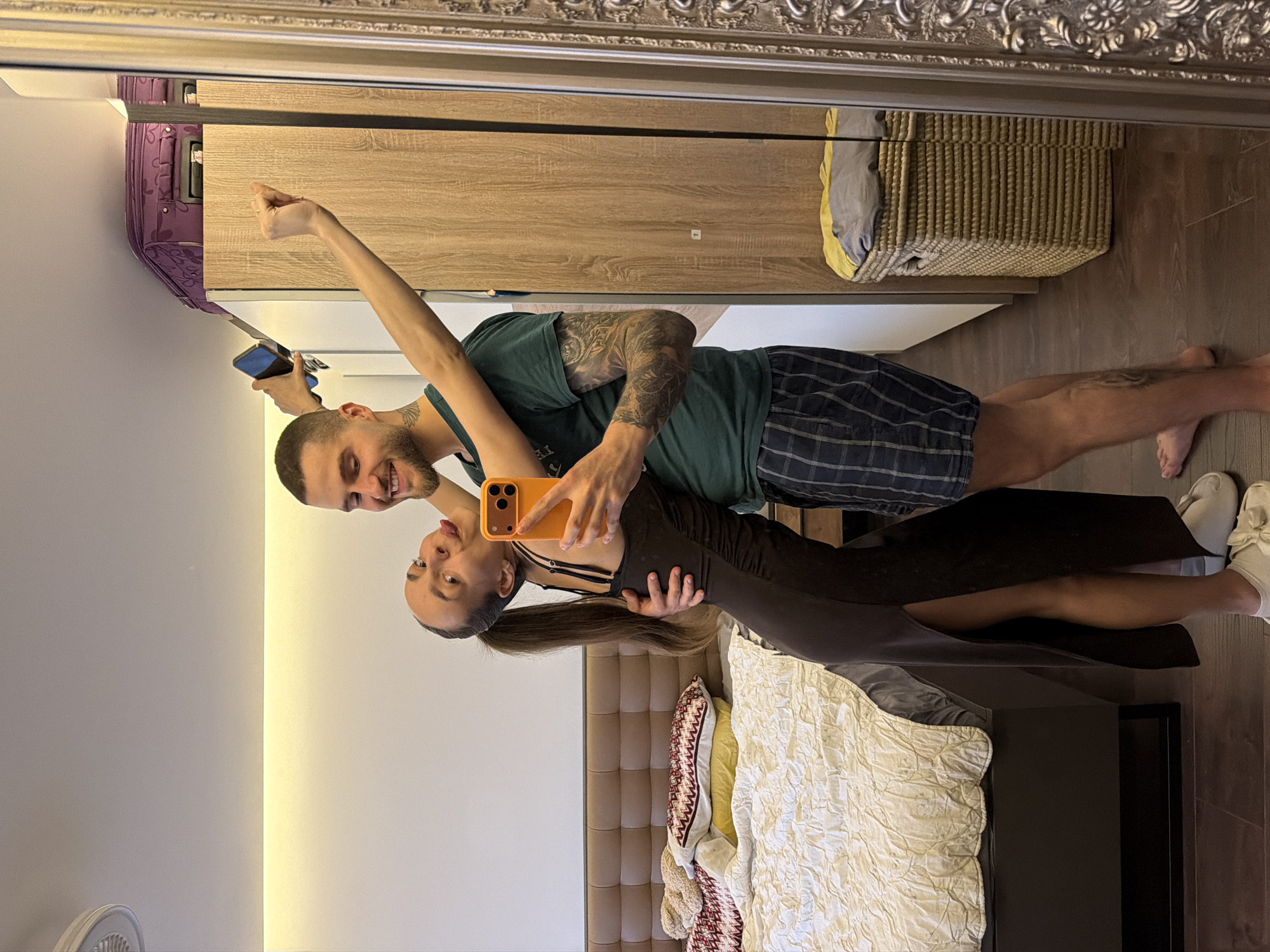 In a bedroom in Sofia, Bulgaria, a man and woman are posing for a selfie in a large, ornate mirror. The woman, wearing a dark, form-fitting dress with a high slit, leans back with her tongue sticking out. She is holding a bright orange smartphone, taking the picture. The man, in a green t-shirt and plaid shorts, has his arm around her. The room features a bed with white bedding and patterned pillows, a wooden wardrobe, and a woven laundry basket. A purple suitcase is visible on top of the wardrobe. The floor is dark wood.