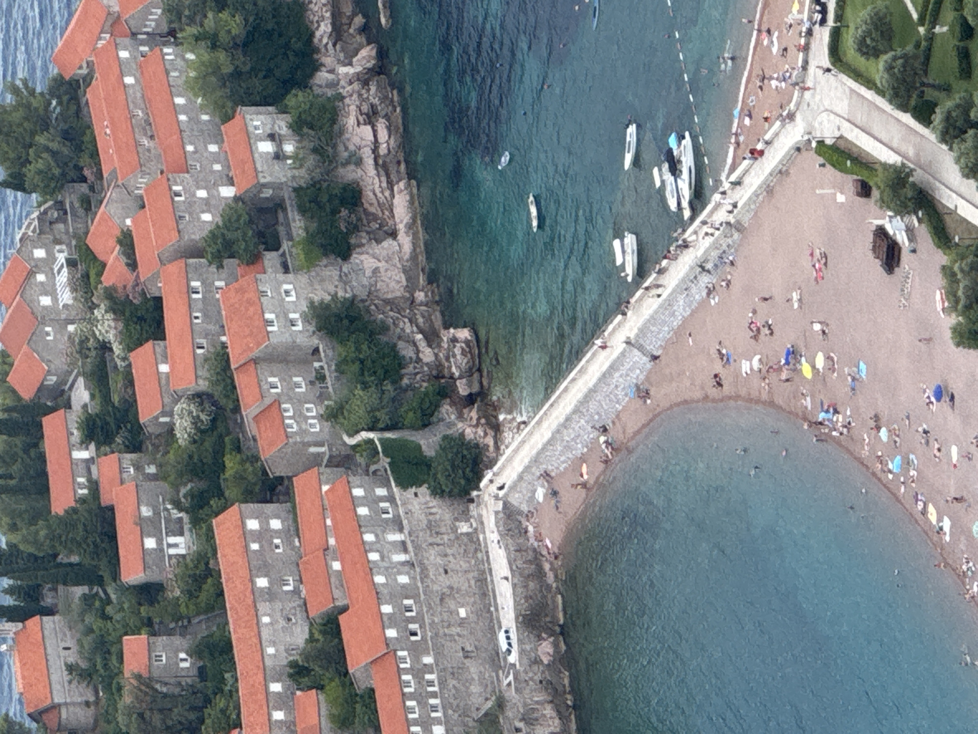 This is an aerial, oblique, tilt-shifted photograph of Sveti Stefan, Montenegro. The image showcases a resort island connected to the mainland by a narrow causeway. On the island, traditional stone buildings with red-tiled roofs are clustered together. Lush green trees are interspersed among the structures.
To the right of the island and causeway is a beach with many sunbathers and umbrellas. The sand is light brown, and the sea is a clear, tranquil turquoise. Several boats are anchored in the water near the island and along the coastline. The activity appears to be a typical summer day at a popular tourist destination, with people relaxing on the beach and enjoying the water.
The scene is bathed in bright daylight, suggesting it's midday or early afternoon. The weather seems fair, with no visible clouds. The distinct architecture of the buildings and the coastal landscape are strong location cues for Sveti Stefan. The overall mood is one of leisure and vacation. No visible text is present in the image.